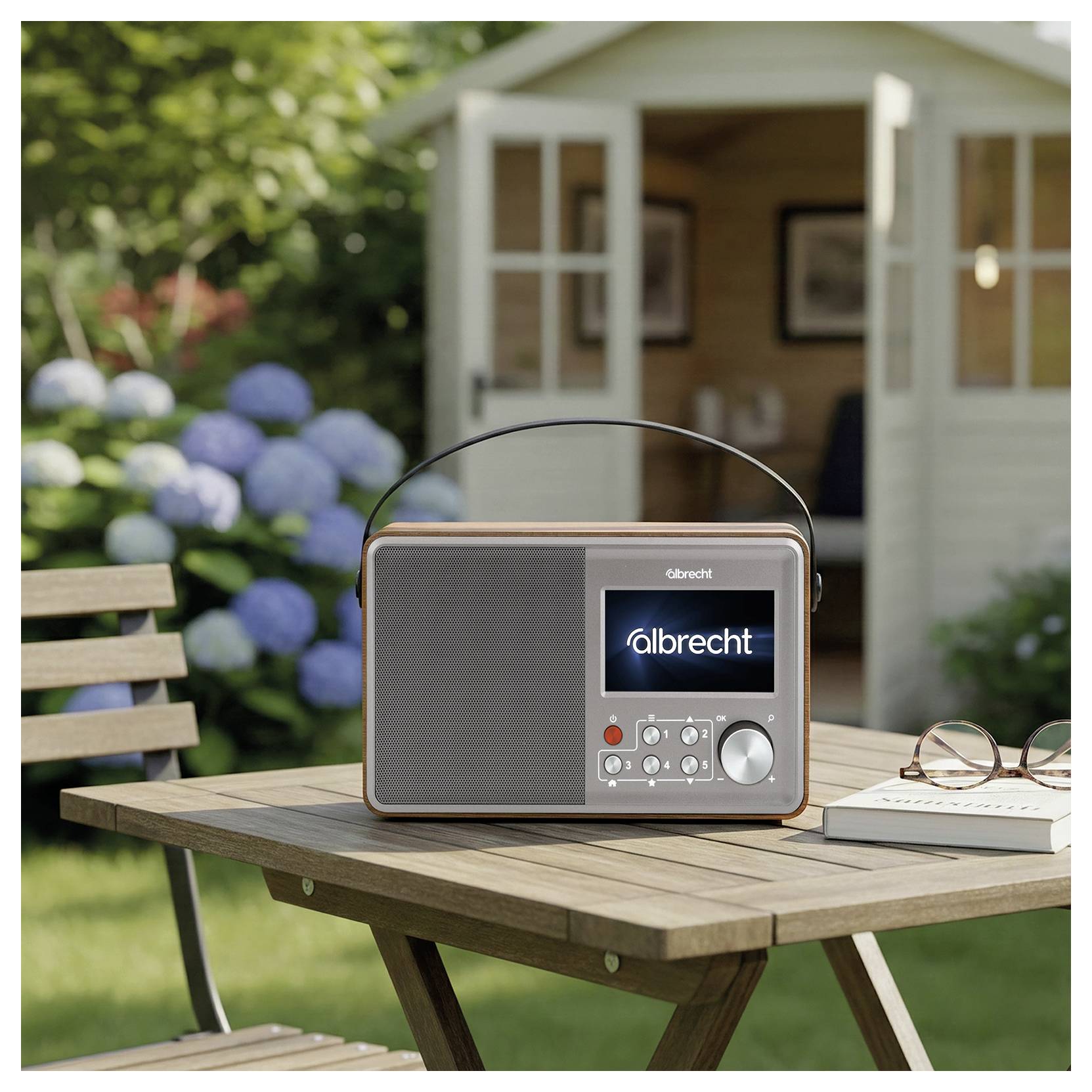 Tragbares Albrecht-Radio auf einem Holztisch im Freien neben einem Buch und einer Brille, mit einem Garten und einem offenen Schuppen im Hintergrund.