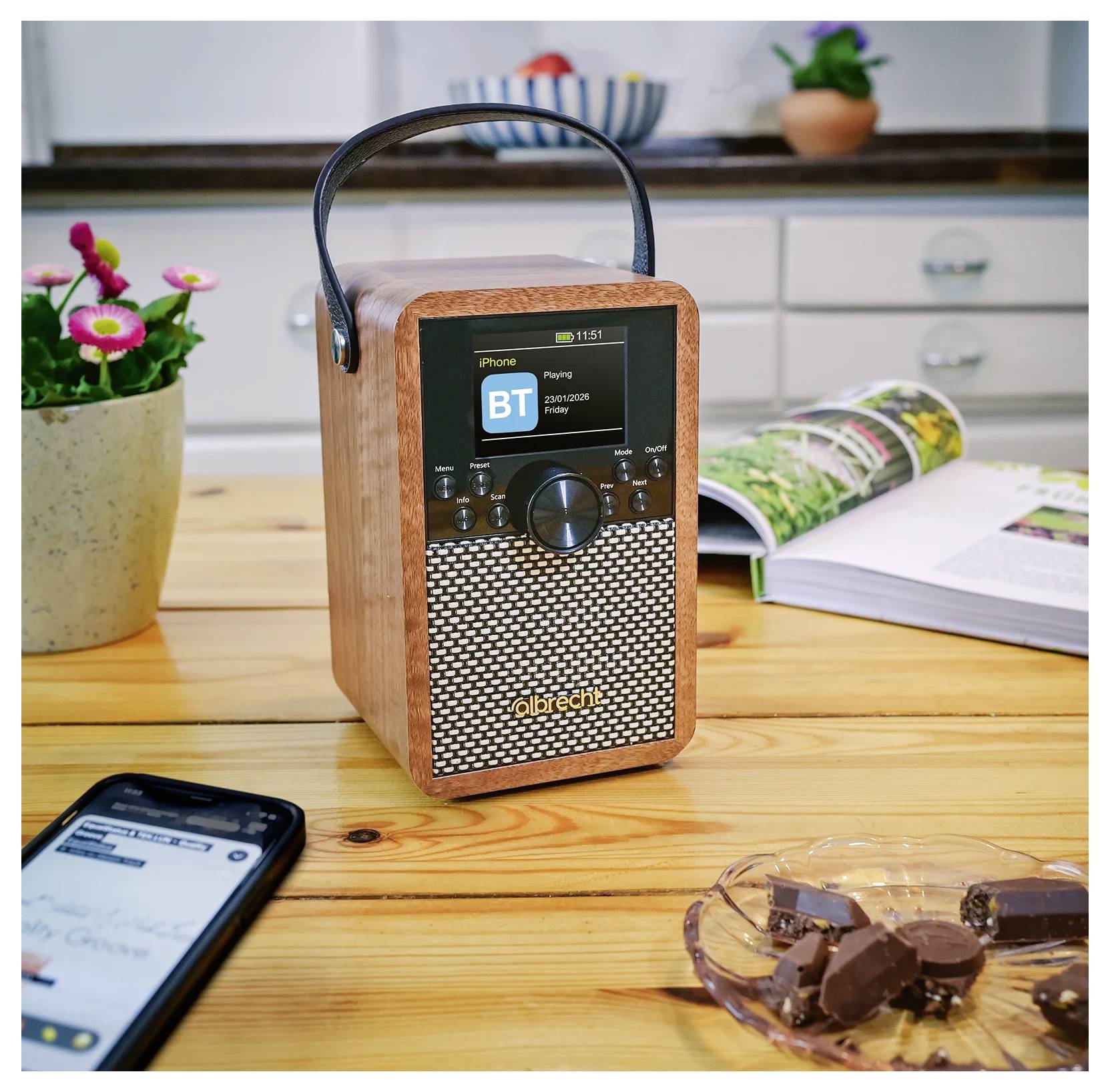 Tragbarer Holzlautsprecher mit Griff auf einem Tisch, der einen Bluetooth-Wiedergabebildschirm anzeigt; Smartphone, aufgeschlagenes Buch, Blumen und Pralinen in der Nähe.