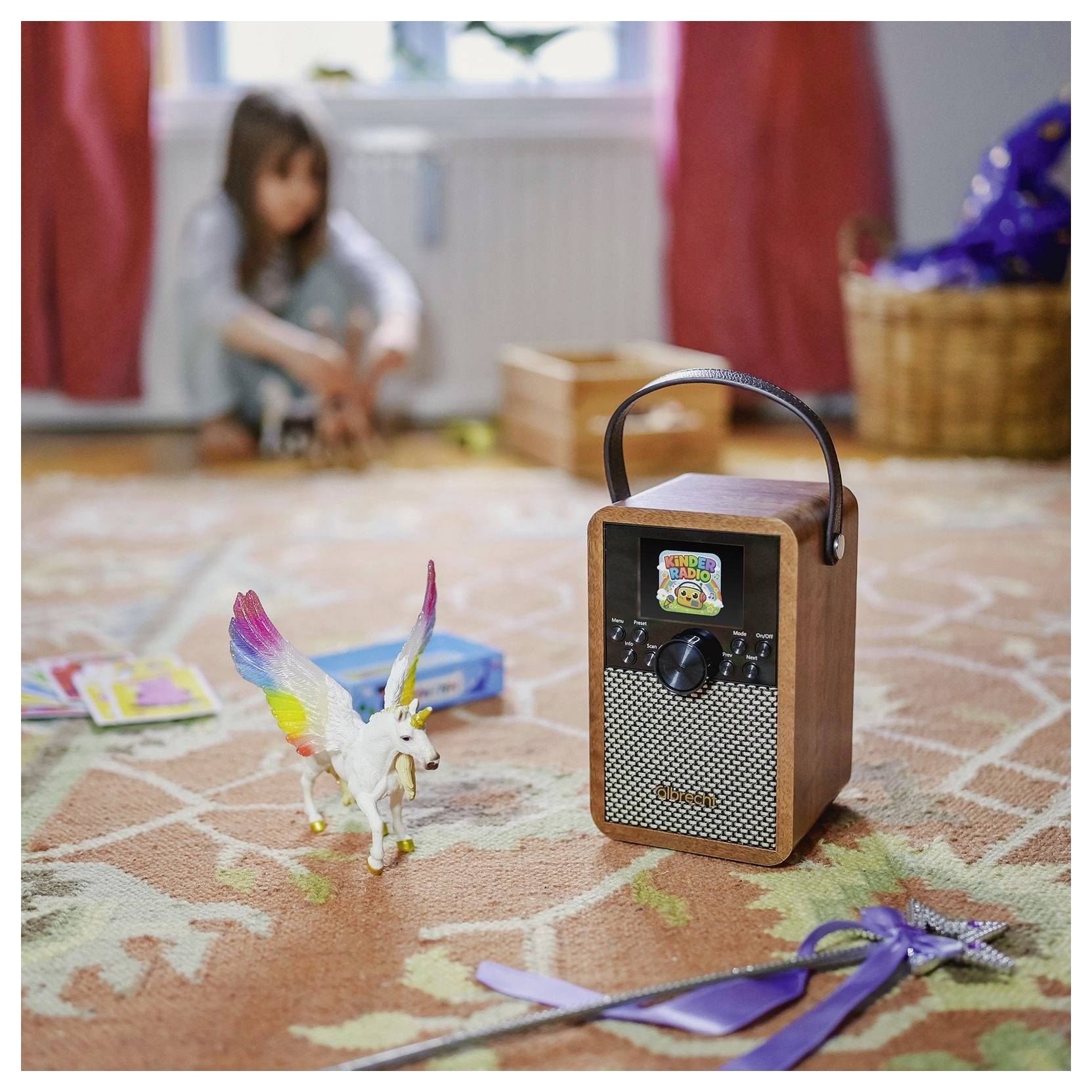Ein Spielzeugradio auf einem gemusterten Teppich neben einer kleinen geflügelten Einhorn-Figur, mit einem verschwommenen Kind im Hintergrund.