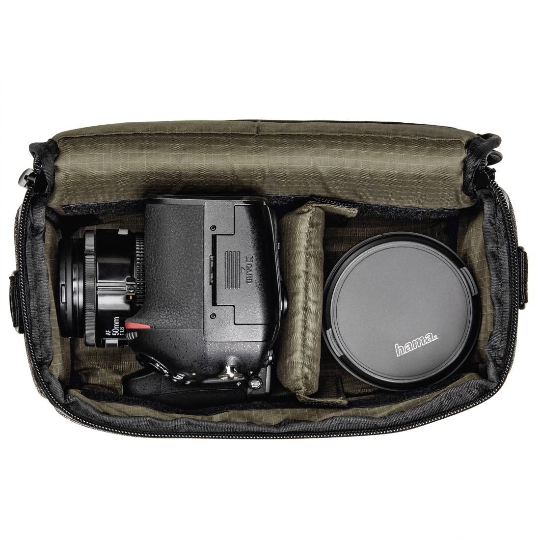 Offene Kameratasche mit einer DSLR-Kamera auf der linken Seite und einem Objektiv mit Kappe auf der rechten Seite, getrennt durch gepolsterte Trennwände.