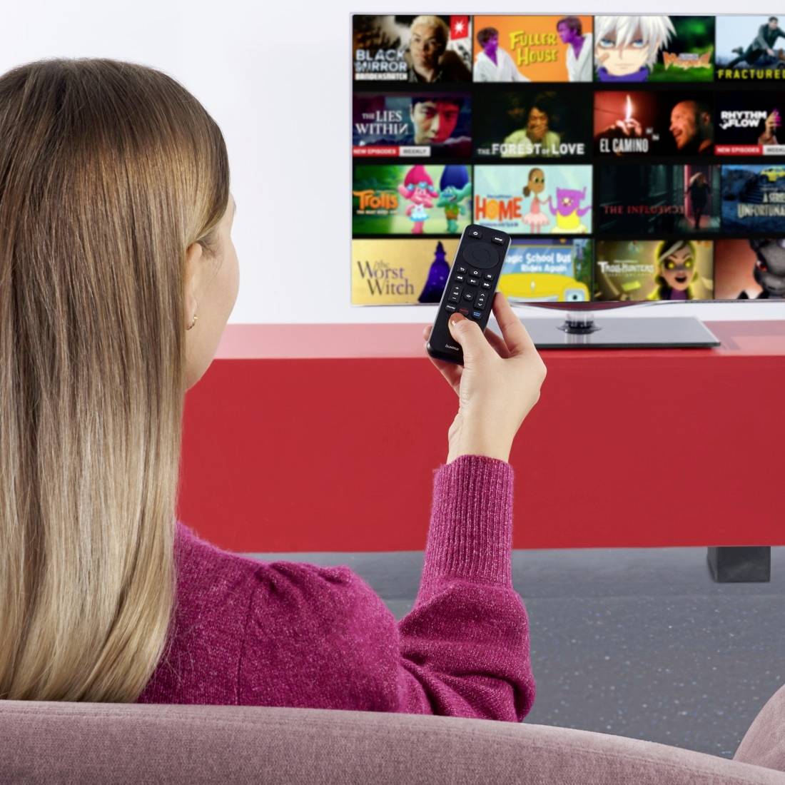 Frau sitzt auf einem Sofa und benutzt eine Fernbedienung, um in einem Streaming-TV-Menü mit Show-Miniaturansichten auf dem Bildschirm zu blättern.