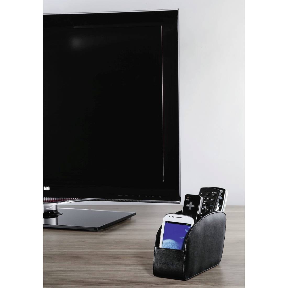Schreibtisch mit großem Monitor und schwarzem Organizer, in dem ein Smartphone und Fernbedienungen auf einer Holzoberfläche aufbewahrt werden.