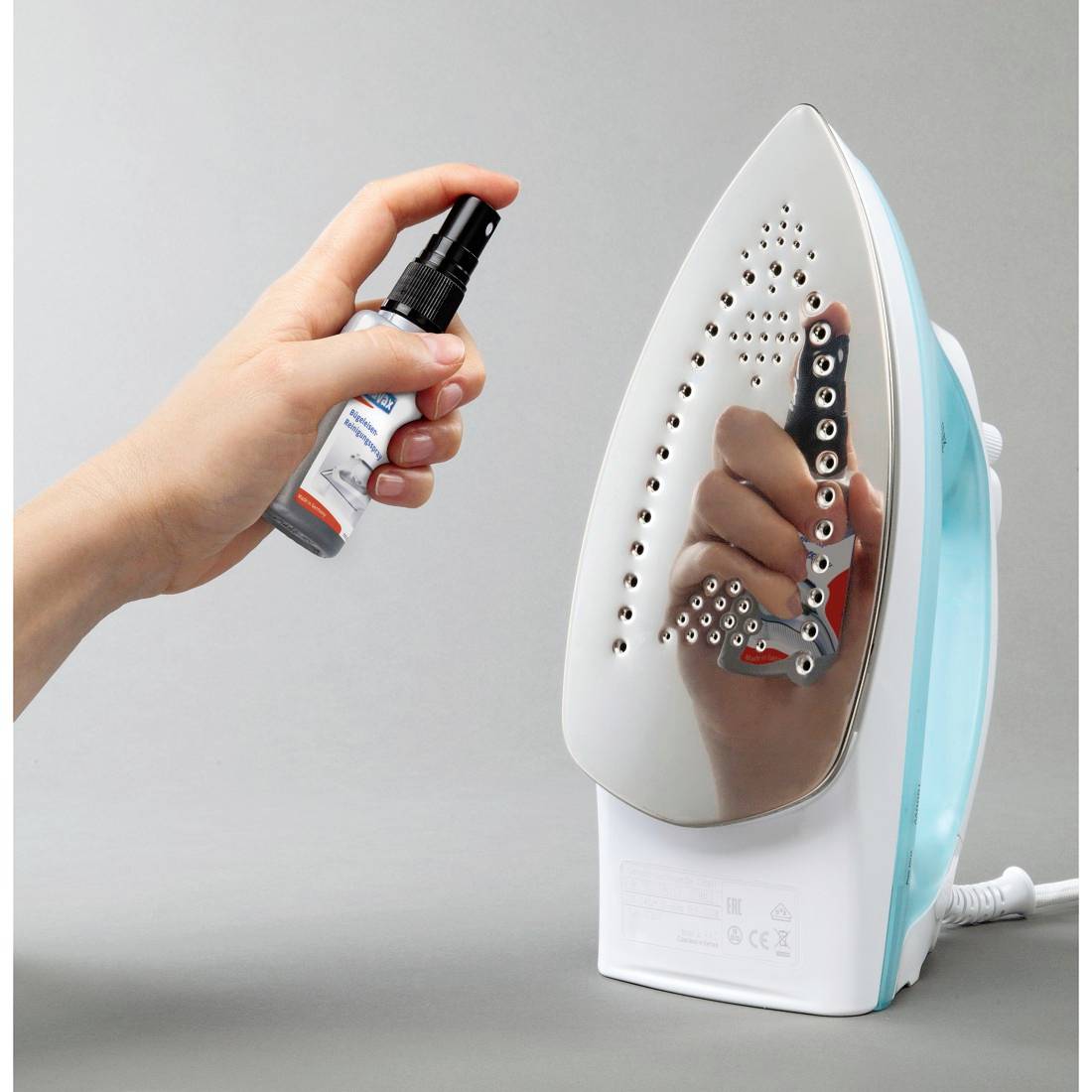 Hand sprays cleaner onto a standing electric iron, suggesting ironing or appliance maintenance.---Hand sprüht Reiniger auf ein stehendes Elektrobügeleisen, was auf Bügeln oder Wartung von Haushaltsgeräten hindeutet.