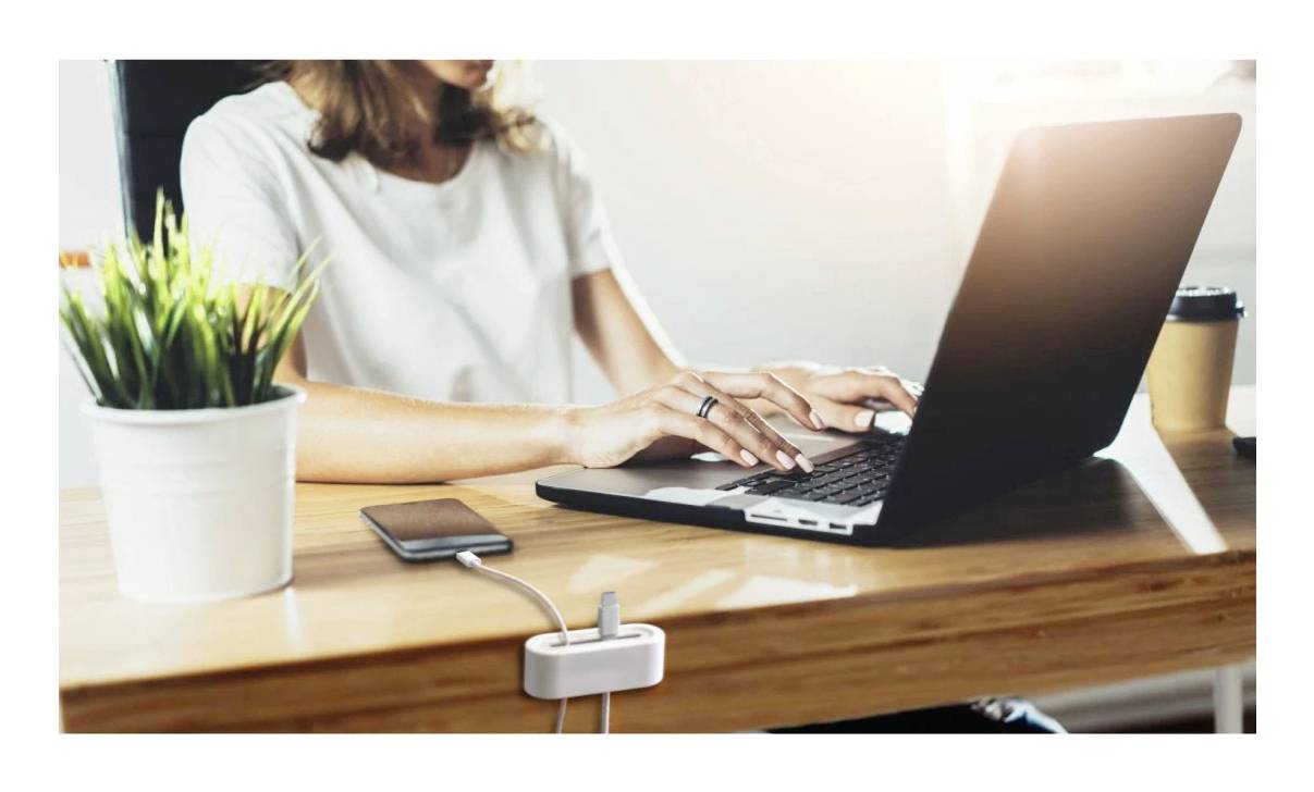 Person tippt auf einem Laptop an einem Holzschreibtisch, wobei ein Smartphone neben einer Topfpflanze und einem Becher zum Mitnehmen aufgeladen wird.