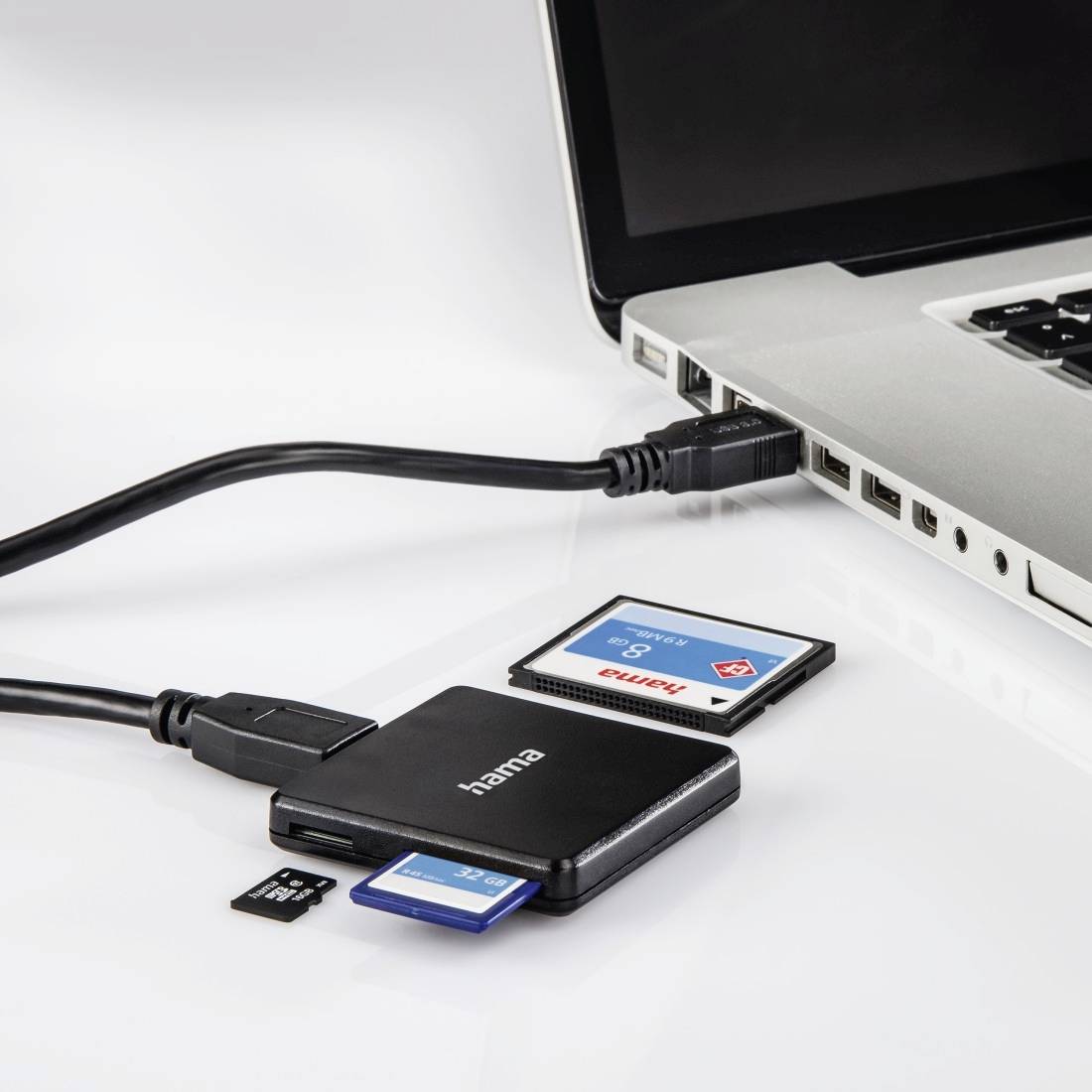 Laptop, das per Kabel mit einem Hama-Kartenleser verbunden ist, mit eingelegten SD- und microSD-Speicherkarten auf einer weißen Oberfläche.