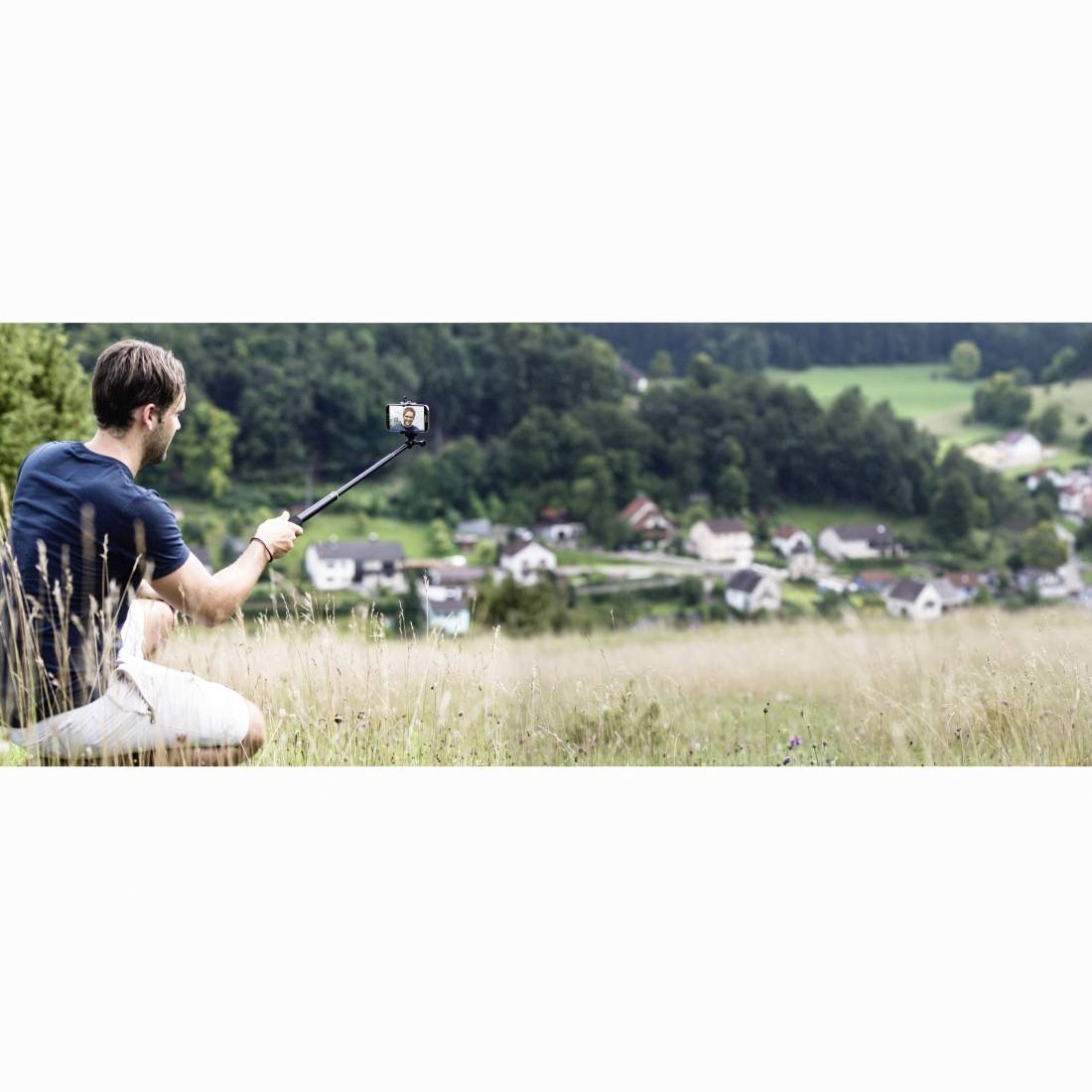 Ein Mann macht ein Selfie mit einem Selfiestab, während er in einem grünen Feld sitzt und auf ein Dorf und bewaldete Hügel blickt.