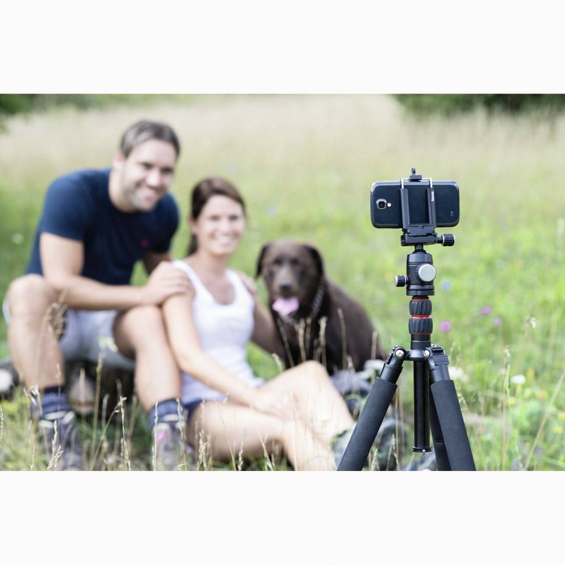 Ein Smartphone auf einem Stativ nimmt ein lachendes Paar und seinen Hund auf, die in einem Grasfeld sitzen.