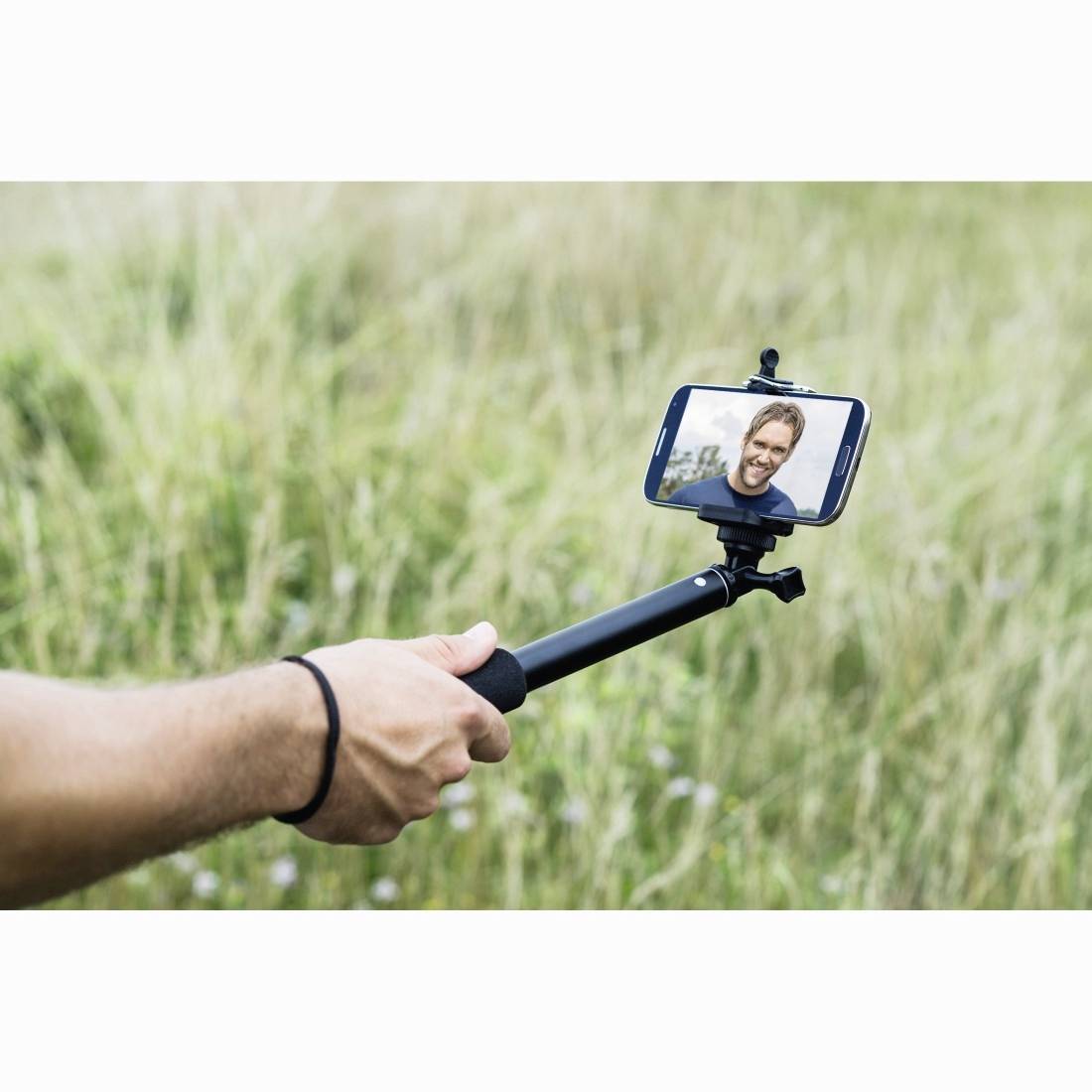 Eine Person, die einen Selfiestab mit einem Smartphone hält, das einen lächelnden Mann auf dem Bildschirm zeigt, draußen im hohen Gras.