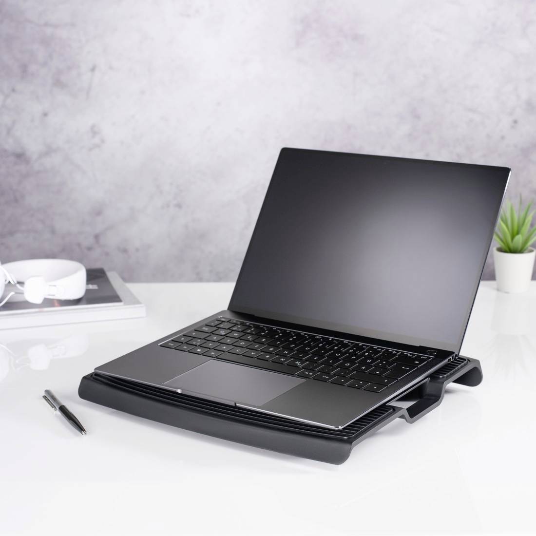 Schwarzer Laptop auf einem Ständer auf einem weißen Schreibtisch, mit einem Stift, Notizbuch, Kopfhörern und einer kleinen Pflanze in einer minimalistischen Büroumgebung.