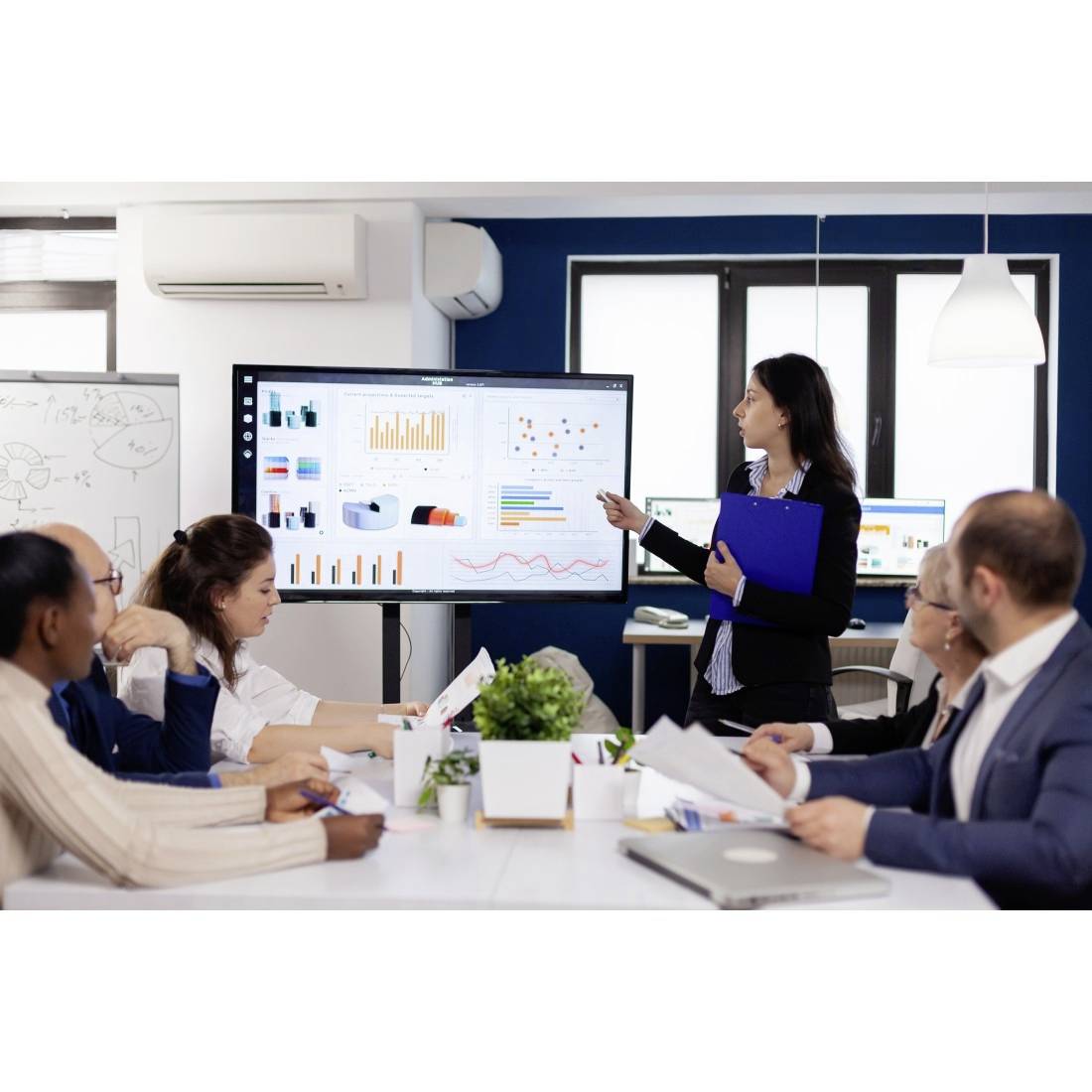 Ein Geschäftspräsentator zeigt auf Dashboard-Diagramme während einer Teambesprechung in einem Konferenzraum, während Kollegen um einen Tisch sitzen und Notizen machen.
