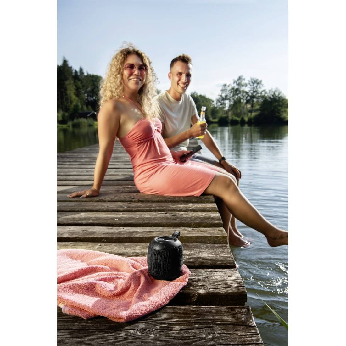 Zwei Menschen entspannen sich auf einem hölzernen Steg an einem See; eine Frau in einem rosa Kleid sitzt vorne, während ein Mann hinter ihr ein Getränk hält.