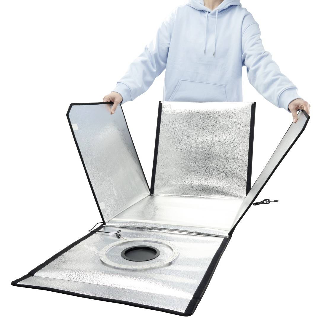 Person in einem hellen Hoodie, der einen reflektierenden zusammenklappbaren Studio-Lichtreflektor mit einer kreisförmigen zentralen Öffnung auf weißem Hintergrund öffnet.