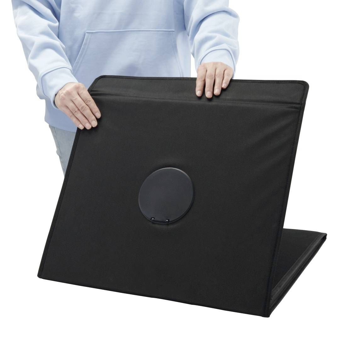 Person in einem hellblauen Hemd, der ein großes schwarzes faltbares Panel mit einer zentralen kreisförmigen Öffnung vor weißem Hintergrund hält.