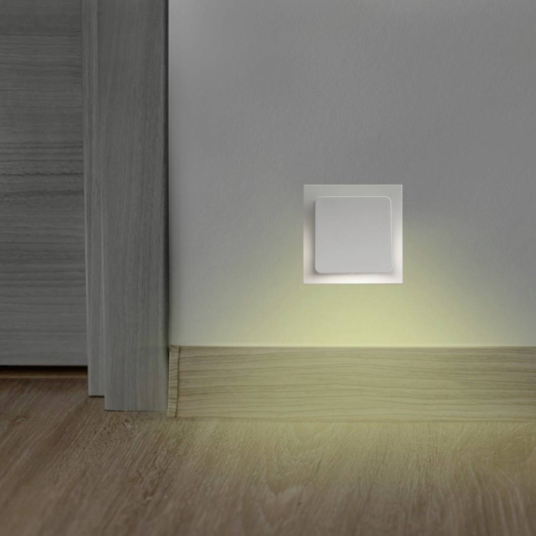 Ein an der Wand montiertes quadratisches Nachtlicht in Bodennähe leuchtet sanft entlang der Fußleiste in einem Flur oder einer Raumecke.