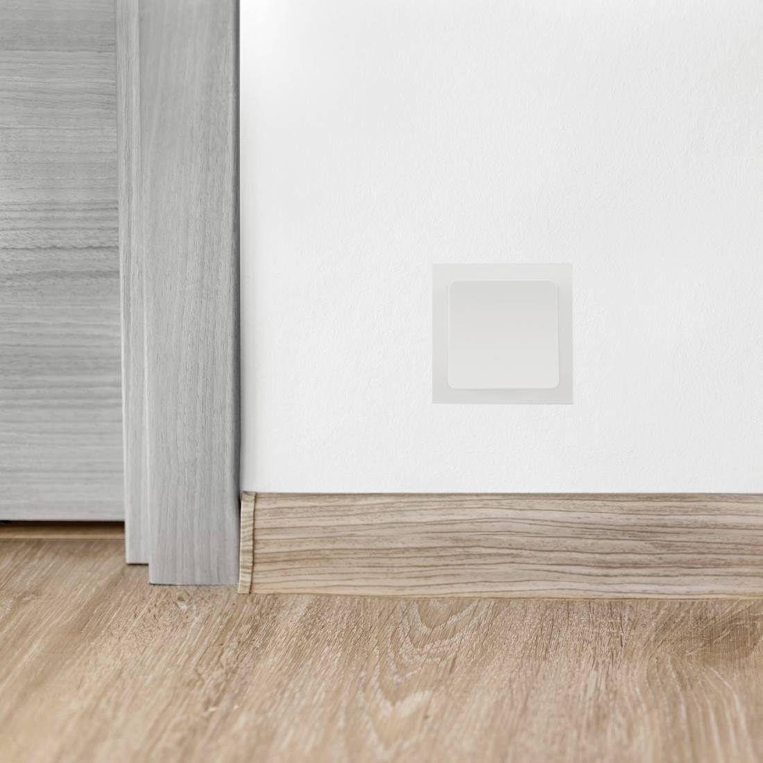 Weiße Wand mit einer leeren quadratischen Wandplatte in Bodennähe neben einem Holztürrahmen und einer Fußleiste auf einem Holzboden.
