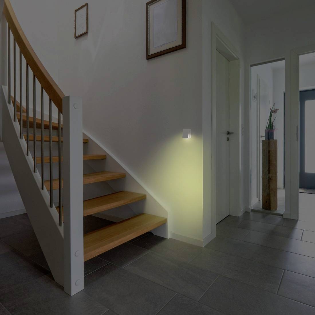 Moderner Flur mit hölzerner Treppe, grauem Fliesenboden und Wandleuchte, die das weiße Interieur in der Nähe einer Türöffnung beleuchtet.
