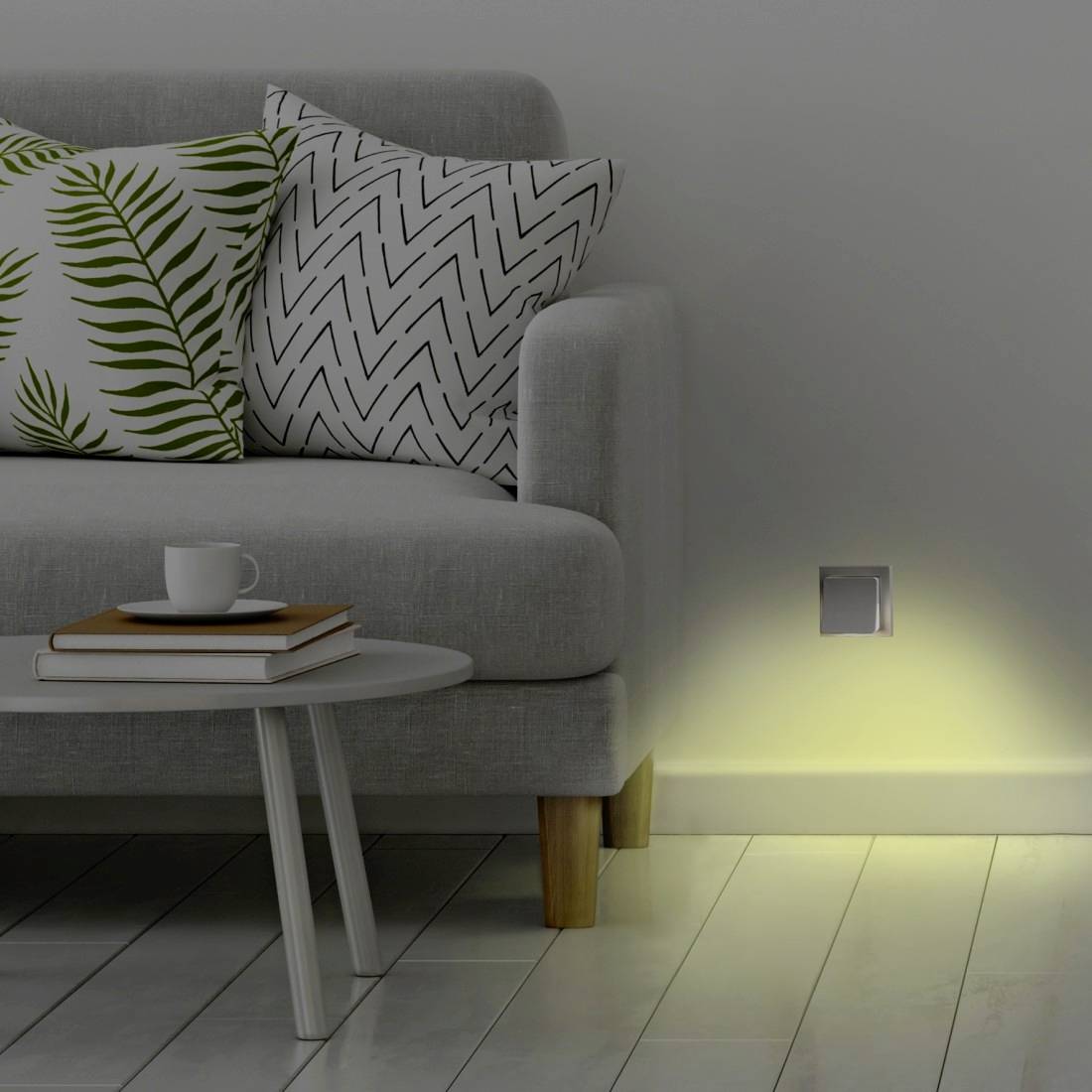Modernes Wohnzimmer mit grauem Sofa, gemusterten Kissen, Beistelltisch mit Büchern und Tasse sowie einer Wandleuchte, die ein sanftes Licht auf den Boden wirft.