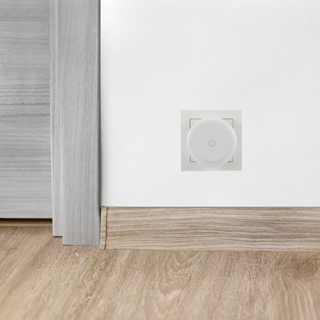 Weiße Wand mit einem kleinen quadratischen Stromschalter/Bedienfeld über einem Holzboden und einer Fußleiste in der Nähe einer Türöffnung oder Schrankecke.