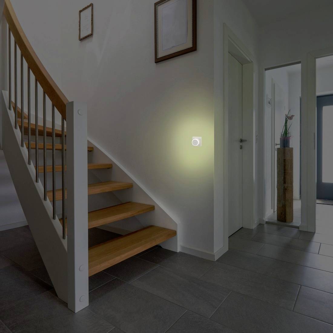 Flur mit hölzerner Treppe und an der Wand montierter Leuchte, die auf einer weißen Wand leuchtet, neben Türen, die zu anderen Räumen führen.
