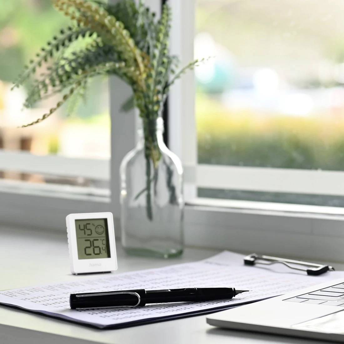 Arbeitsplatz neben einem Fenster mit einem digitalen Thermometer, das 45° und 26° anzeigt, Papiere mit einem Stift, eine Ecke eines Laptops und eine Vase mit Grünpflanzen.