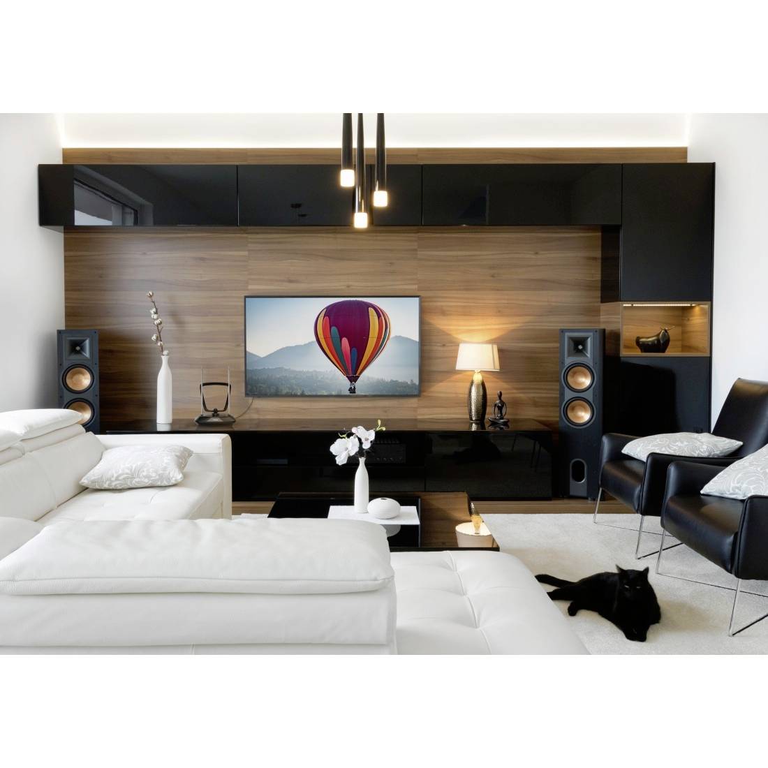 Modernes Wohnzimmer mit weißen Sofas, schwarzen Stühlen, Medienwand aus Holz, Lautsprechern und einem Fernseher, der einen Heißluftballon über Bergen zeigt.