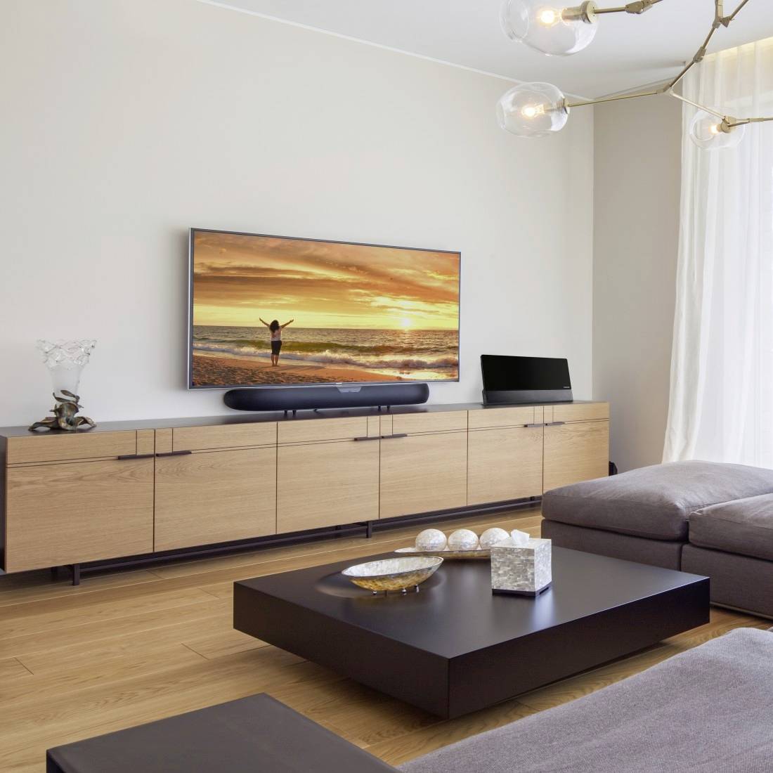 Modernes Wohnzimmer mit an der Wand montiertem Fernseher, der eine Sonnenuntergang-Strandszene zeigt, niedrigem Holzschrank, dunklem Couchtisch, Ecksofa und Pendelleuchten.