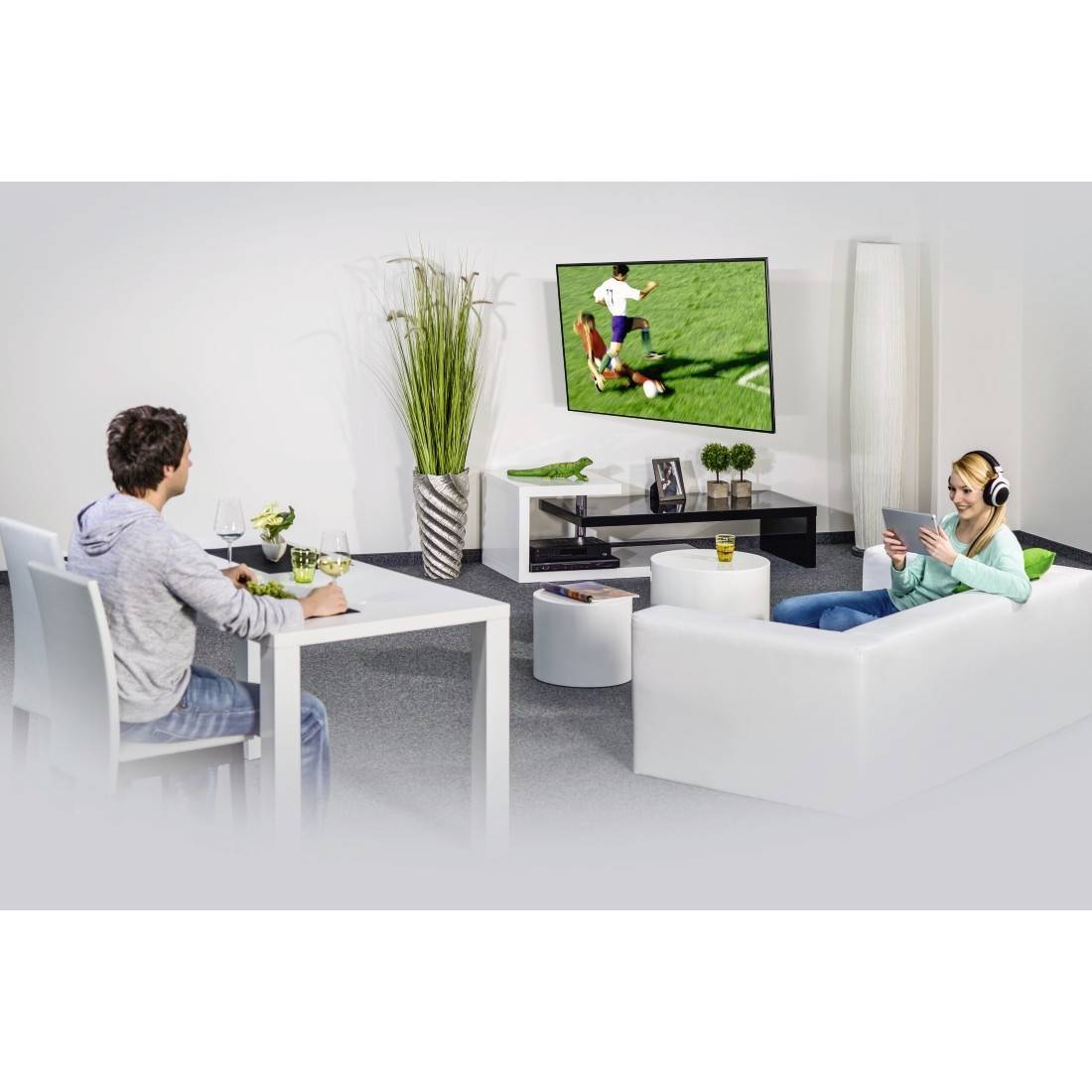 Zwei Personen entspannen sich in einem Wohnzimmer und schauen sich ein Fußballspiel im Fernsehen an; eine sitzt an einem Tisch, die andere liegt auf einem Sofa mit Kopfhörern und einem Tablet.