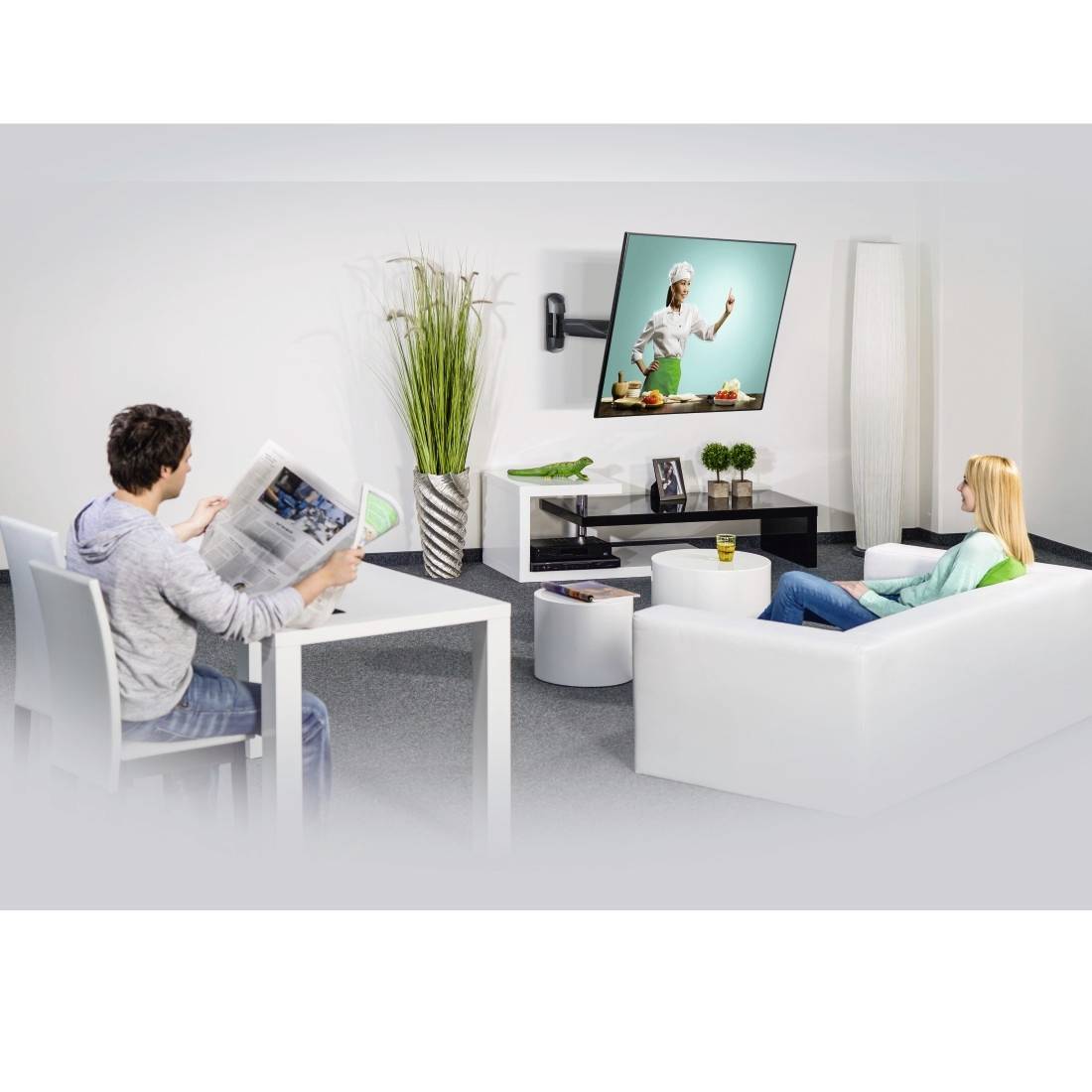 Ein Mann liest eine Zeitung, während eine Frau Fernsehen schaut, in einem modernen Wohnzimmer mit weißen Möbeln und Pflanzen.