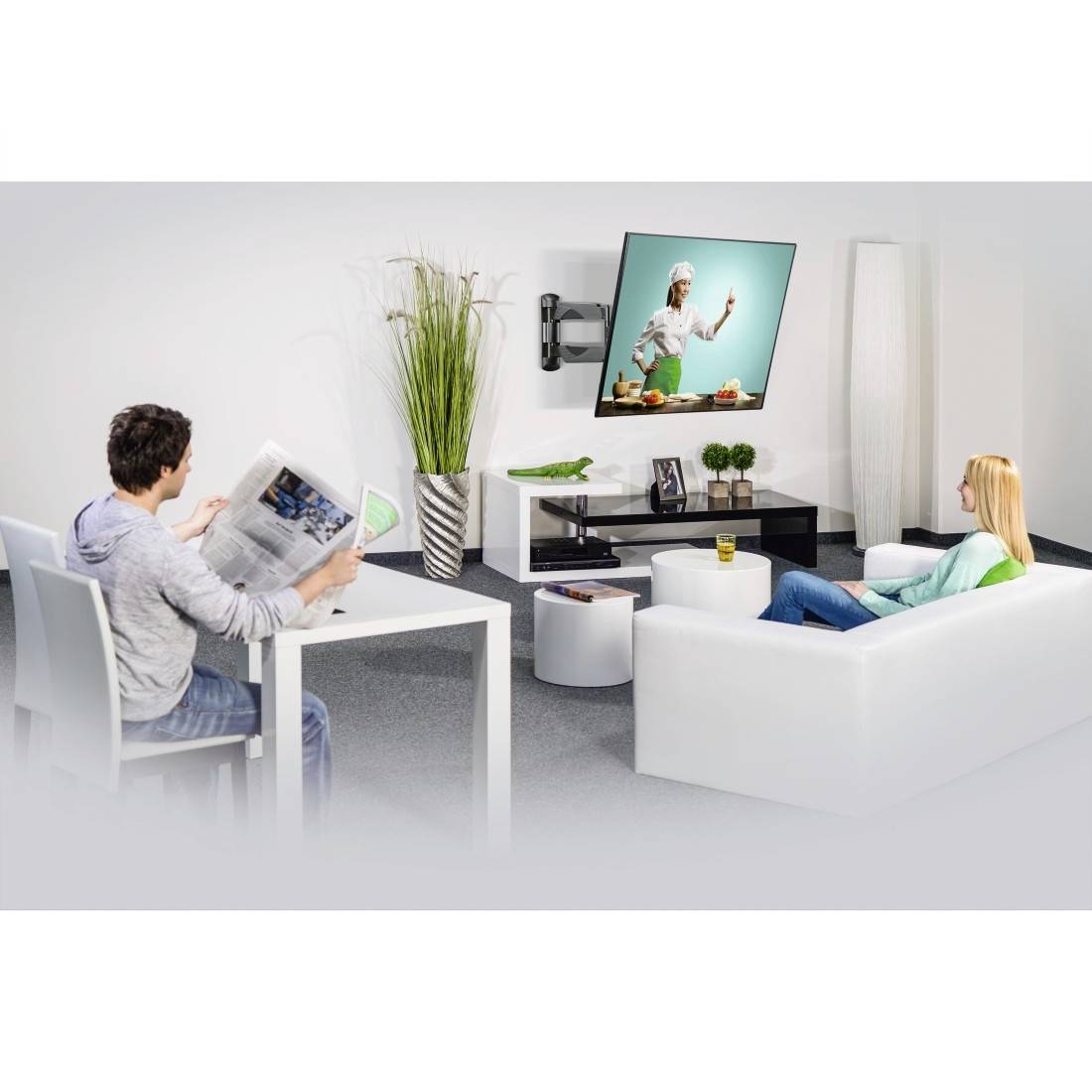 Ein Mann liest eine Zeitung an einem Tisch, während eine Frau in einem modernen Wohnzimmer mit weißen Möbeln fernsieht.