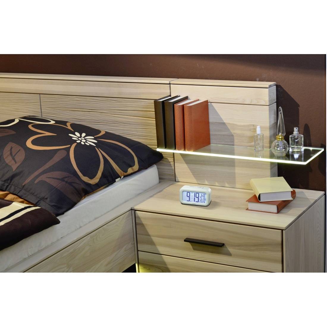 Ein Nachttisch im Schlafzimmer mit einer digitalen Uhr, Büchern, Parfümflaschen und einem Glasregal neben einem Bett mit gemusterten Kissen.