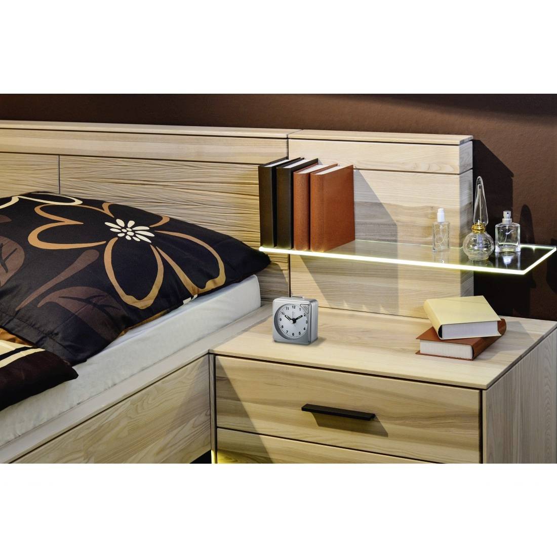Moderner Nachttisch neben einem Bett mit Büchern, einem Wecker, einem kleinen Regal und Glasperfumflaschen auf dem Kopfteil.