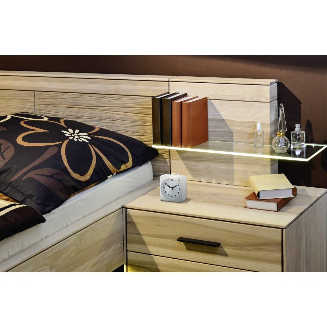 Moderner Schlafzimmerbereich am Bett mit Holzbett, Blumenkissen, Nachttisch, Lampenregal, Wecker, Bücher und kleinen Dekoflaschen.