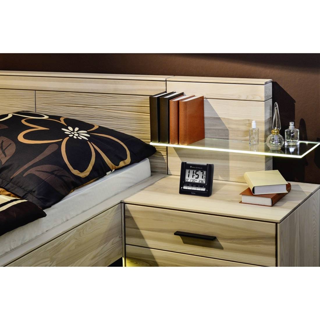 Holztische mit Regal neben dem Bett, Bücher, Uhr zeigt 1:15 Uhr, und Parfumflaschen neben einem Bett mit gemustertem Kissen.