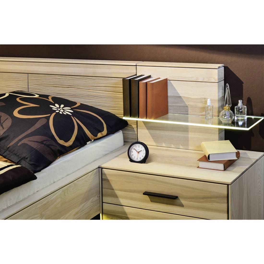 Moderne Schlafzimmerecke mit einem Bett, Nachttisch, Büchern, einer kleinen Uhr und einem Glasregal, das Bücher und Parfümflaschen hält.