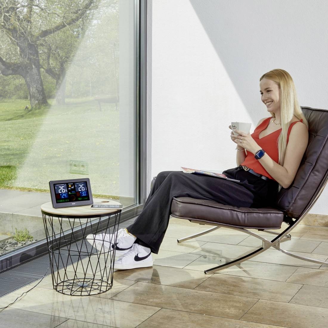 Frau, die sich in einem modernen Stuhl entspannt, hält eine Tasse und liest, neben einem kleinen Tisch mit einer digitalen Wetteruhr neben einem großen Fenster mit Blick auf einen grünen Rasen.