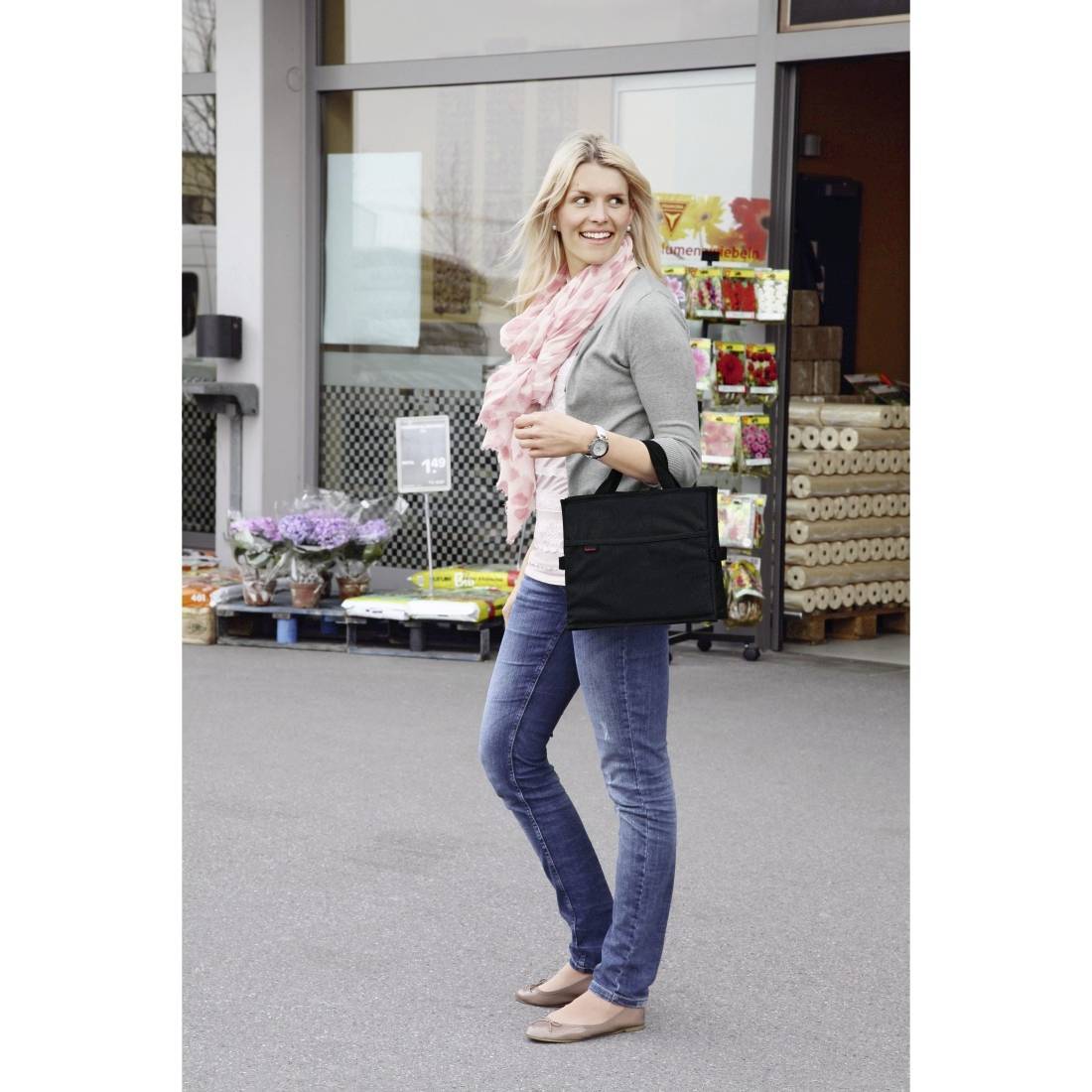 Blonde Frau in einem grauen Pullover, rosa Schal und Jeans steht außerhalb eines Geschäfts und hält eine schwarze Tragetasche.
