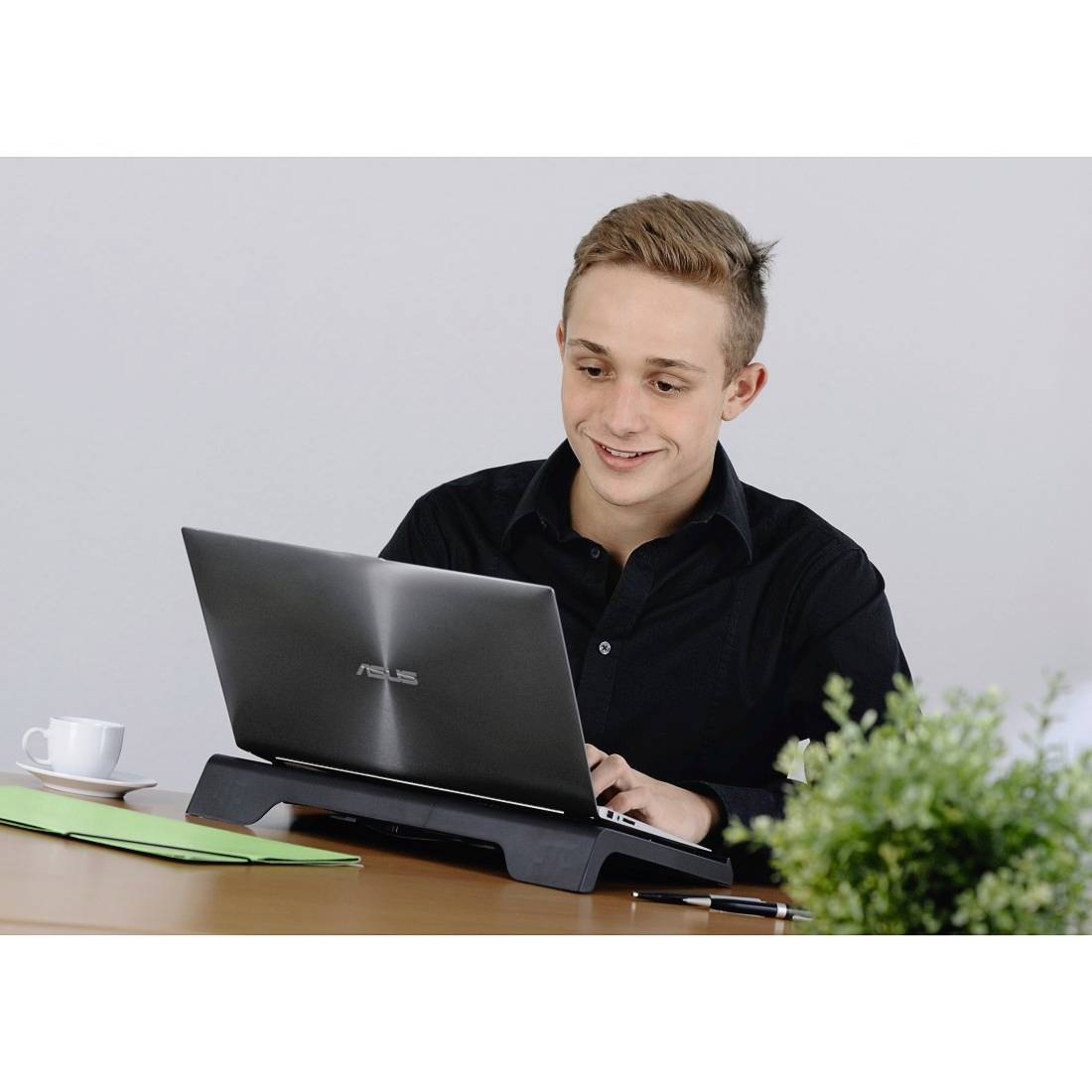 Ein junger Mann in einem schwarzen Hemd lächelt, während er an einem Laptop an einem Schreibtisch mit Papieren, einem Stift und einer Tasse in der Nähe arbeitet.