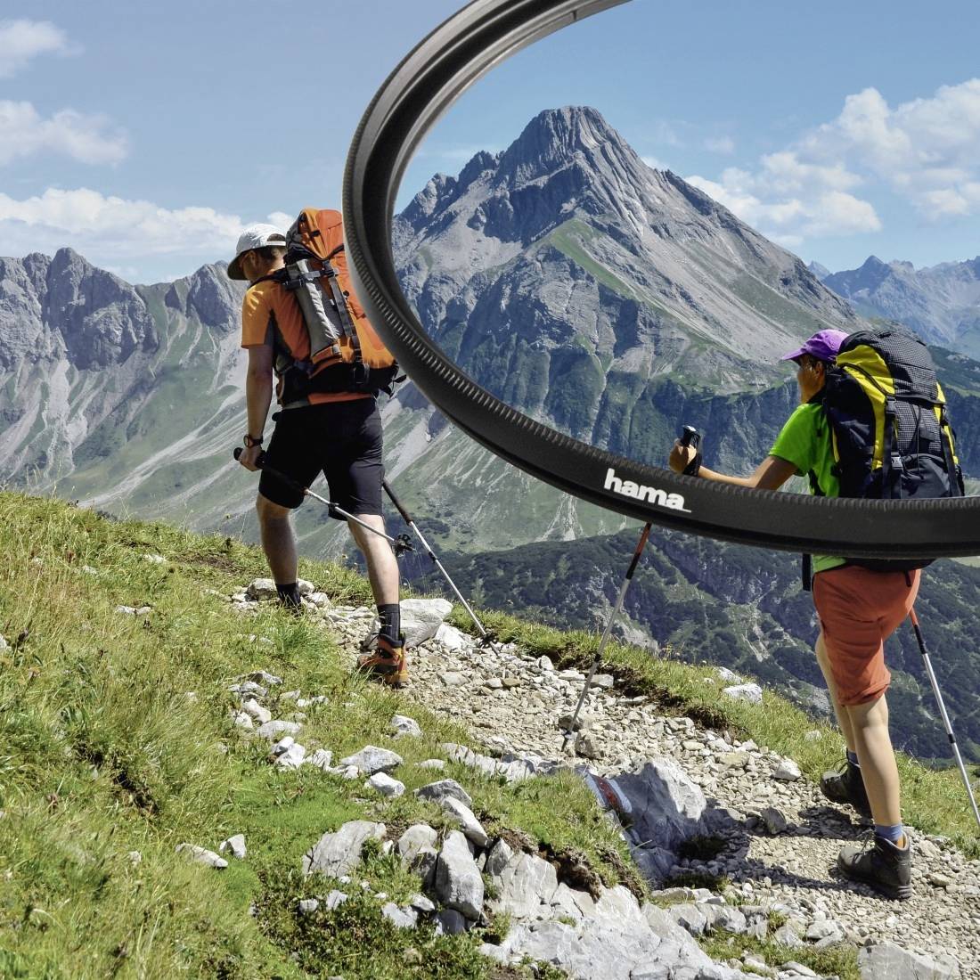 Zwei Wanderer mit Rucksäcken und Trekkingstöcken gehen einen Bergpfad entlang, eingerahmt von einem großen kreisförmigen Objektivfilter, mit dramatischen Gipfeln im Hintergrund.