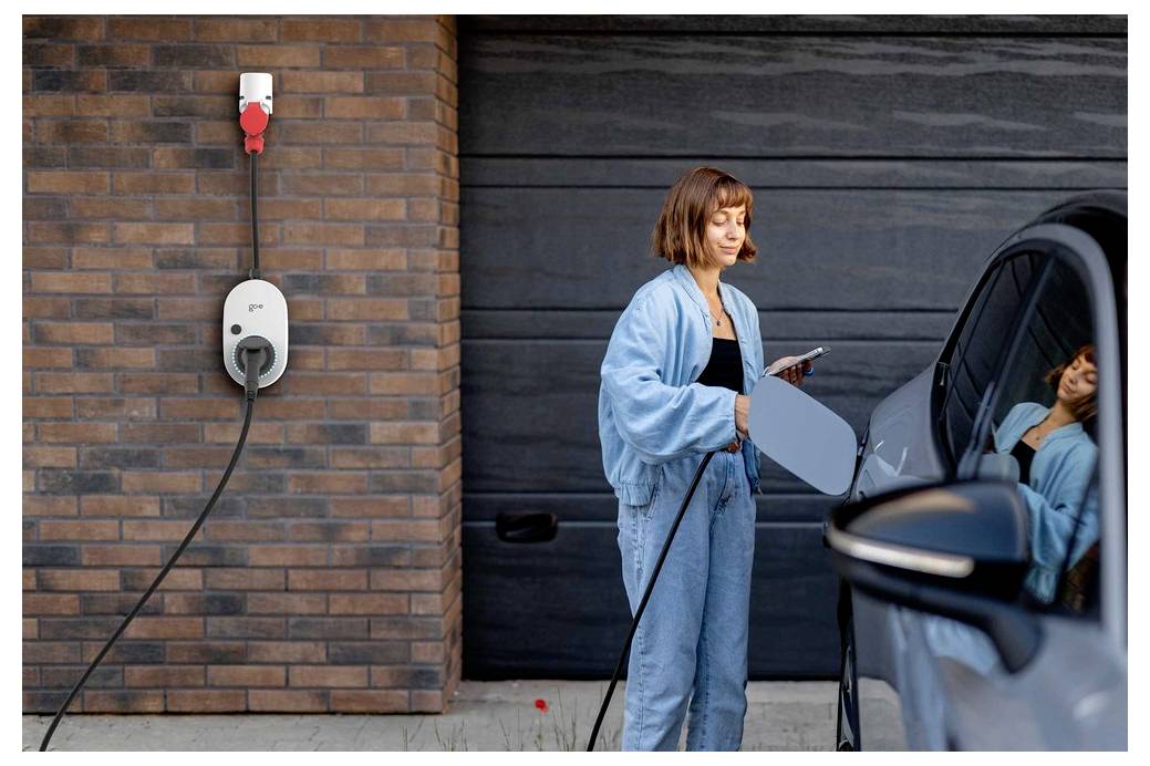 Frau lädt ein Elektroauto auf der heimischen Auffahrt auf und hält ein Telefon neben einer an der Wand montierten Ladestation.