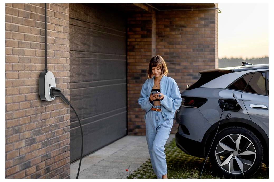 Frau, die ein Smartphone benutzt, während sie neben einem Elektroauto steht, das an einer Wandladestelle außerhalb einer Backsteingarage angeschlossen ist.