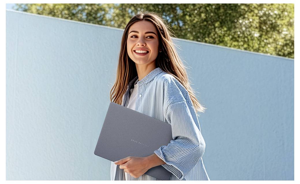 Lächelnde junge Frau im Freien, die einen geschlossenen grauen Laptop gegen eine hellblaue Wand hält.