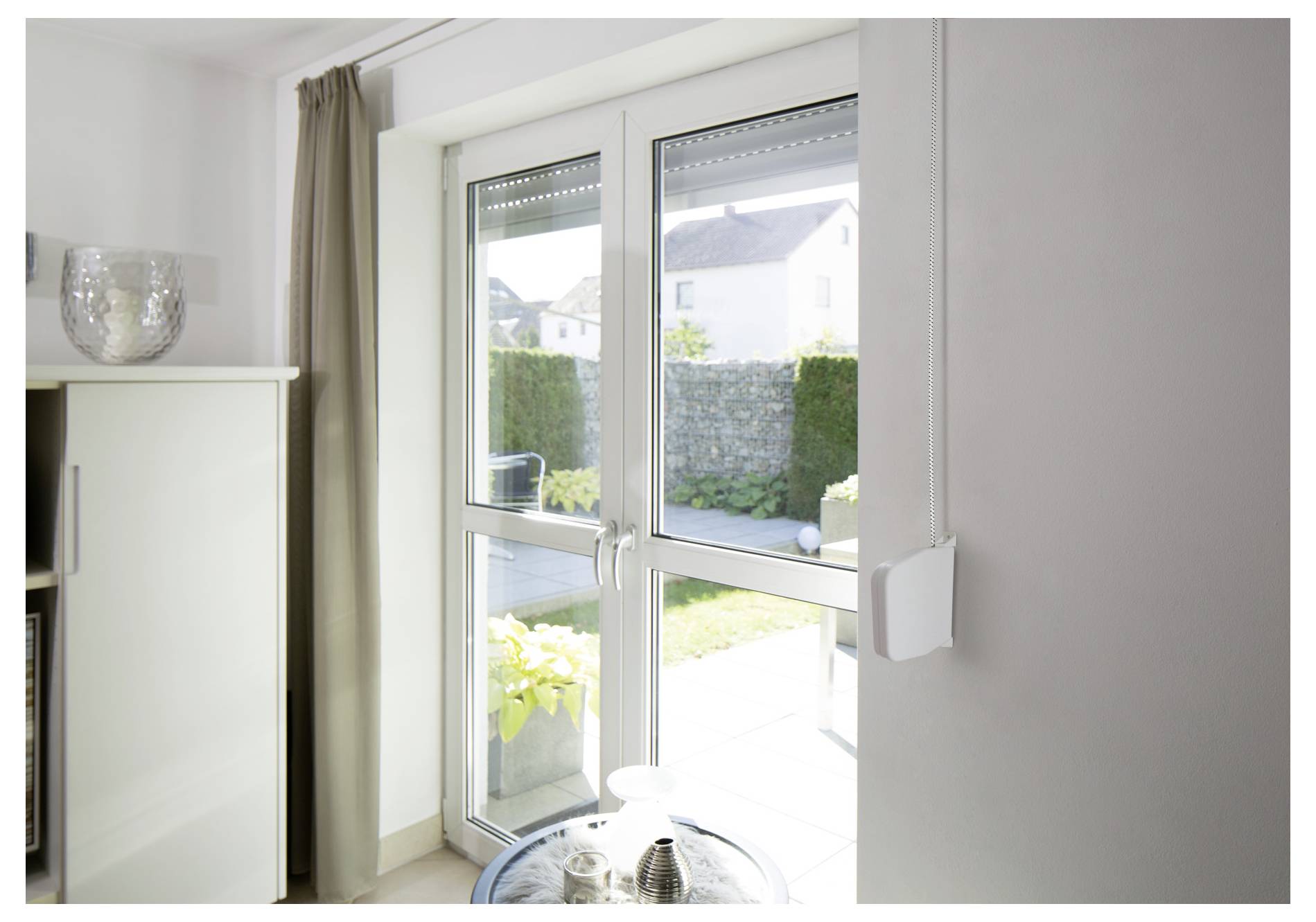 Helles Wohnzimmerinterieur mit einer Glastür und einem Fenster, die sich zu einer sonnigen Terrasse und einem Garten öffnen, mit einem Schrank und Vorhängen auf der linken Seite.