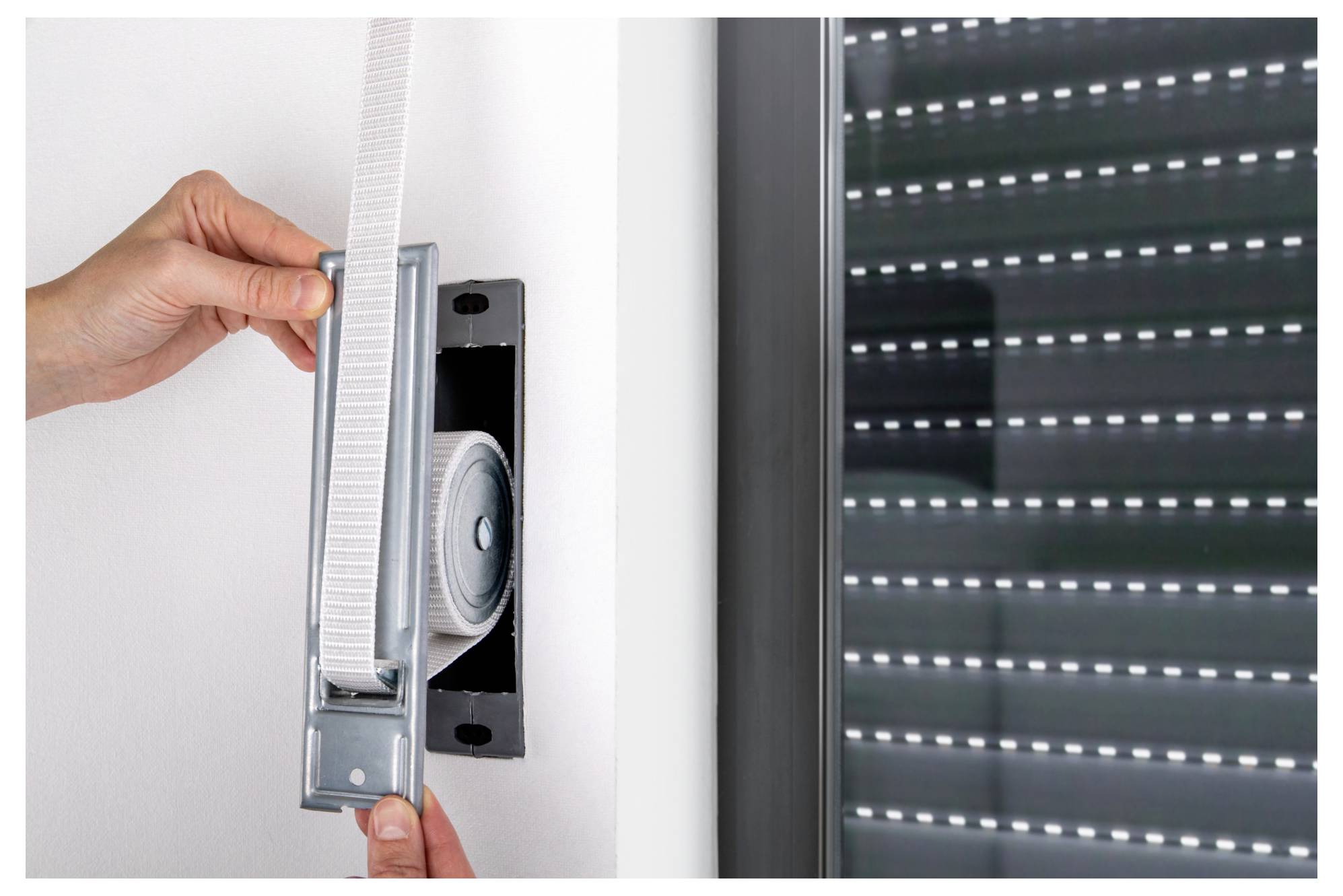 Manuelle Installation eines Metallkettengetriebemechanismus für Fensterjalousien in einer Wandhalterung neben einer geschlossenen Rollojalousie.