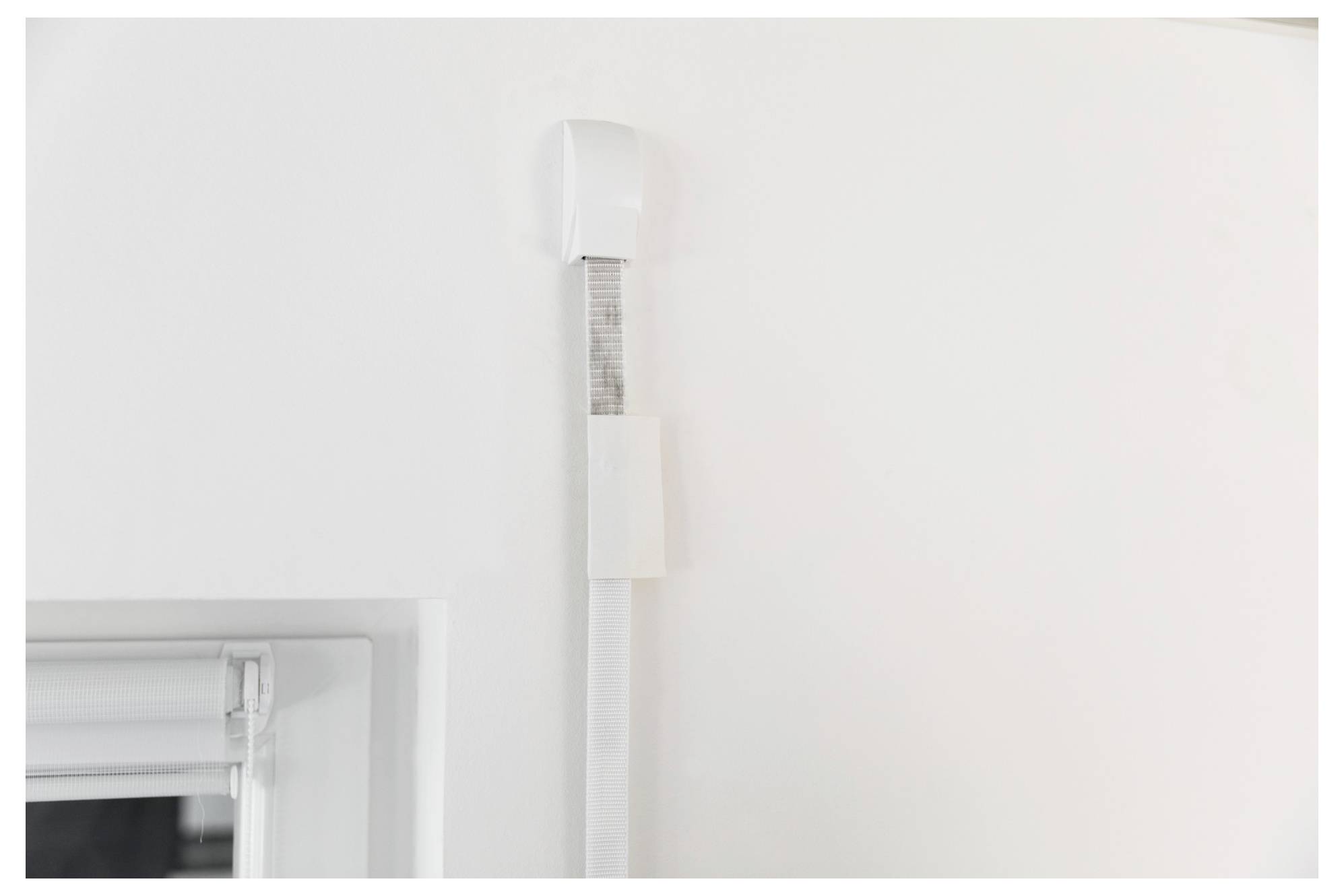 Nahaufnahme eines weißen, an der Wand montierten Kabelkanals oder Sensor-Gehäuses neben einem Fensterrahmen auf einer einfachen weißen Wand.