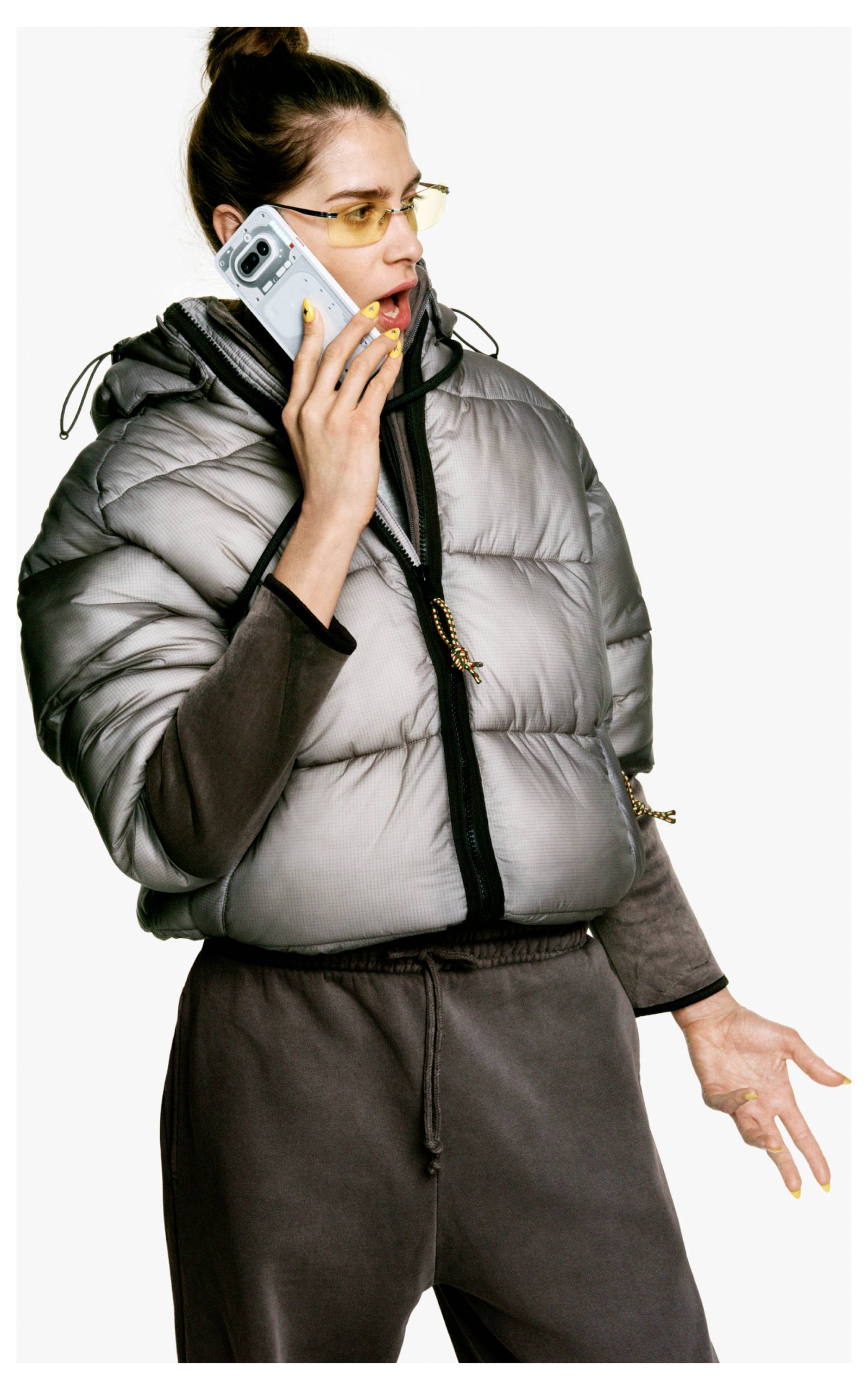 Frau in einer grauen Steppweste und Brille, die ein Smartphone ans Ohr hält und überrascht gegen einen einfachen Hintergrund schaut.