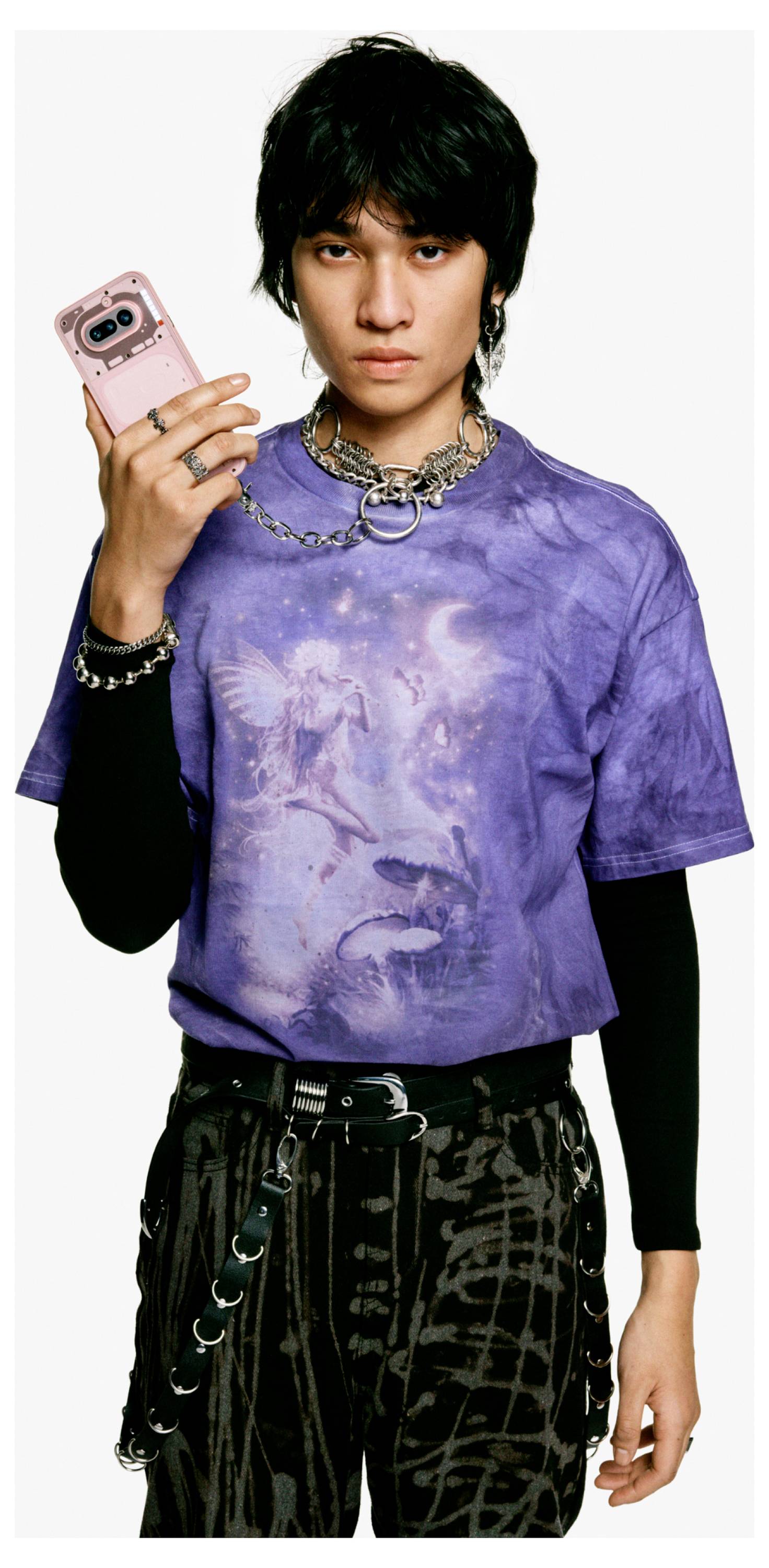 Junger Mann in einem violetten bedruckten T-Shirt und gestuften Ketten hält ein rosa Telefon hoch, posiert vor einem schlichten weißen Hintergrund.
