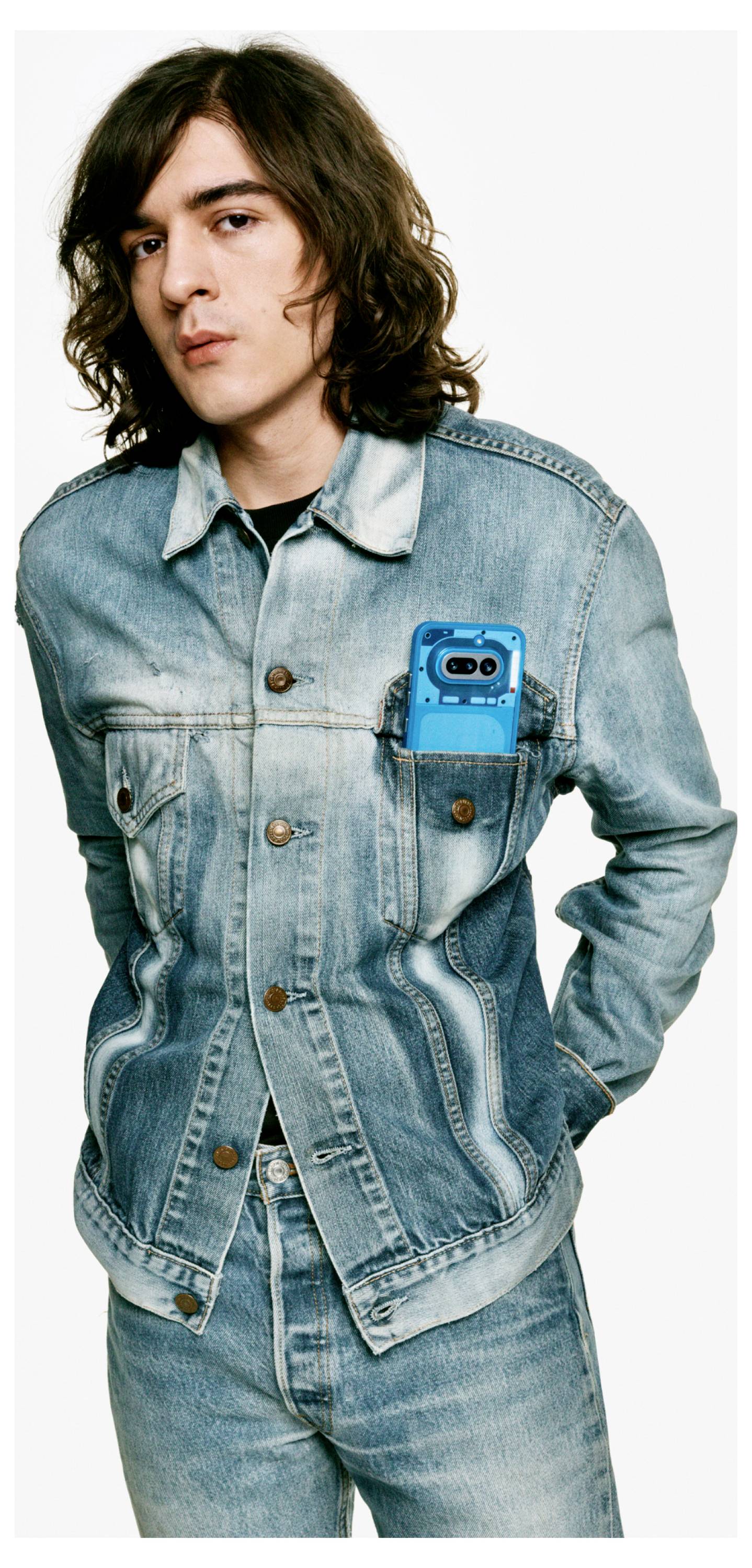 Porträt einer Person in einer hellen Denim-Jacke und Jeans, Hände in den Taschen, mit einem leuchtend blauen Telefon, das in der Brusttasche sichtbar ist.