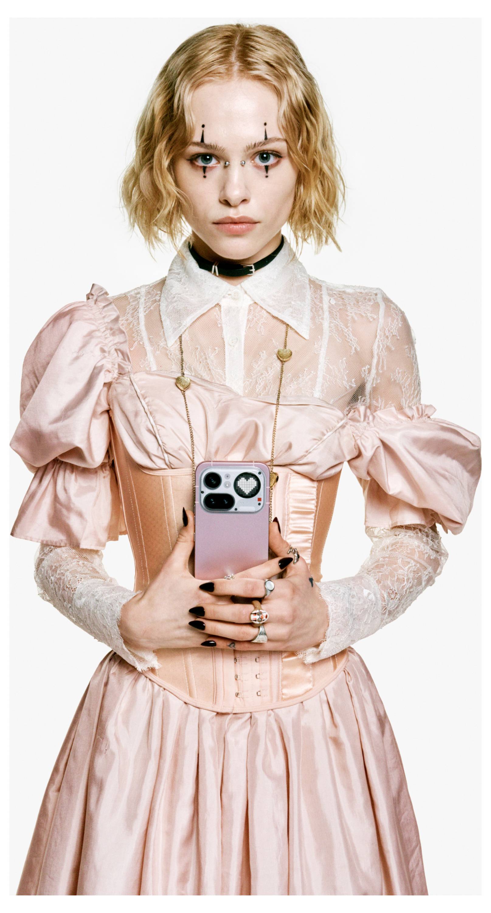 Porträt einer jungen Frau in einem hellrosa Vintage-Kleid, die ein Smartphone hält, mit goldenem Schmuck und Gesichtsjuwelen vor weißem Hintergrund.