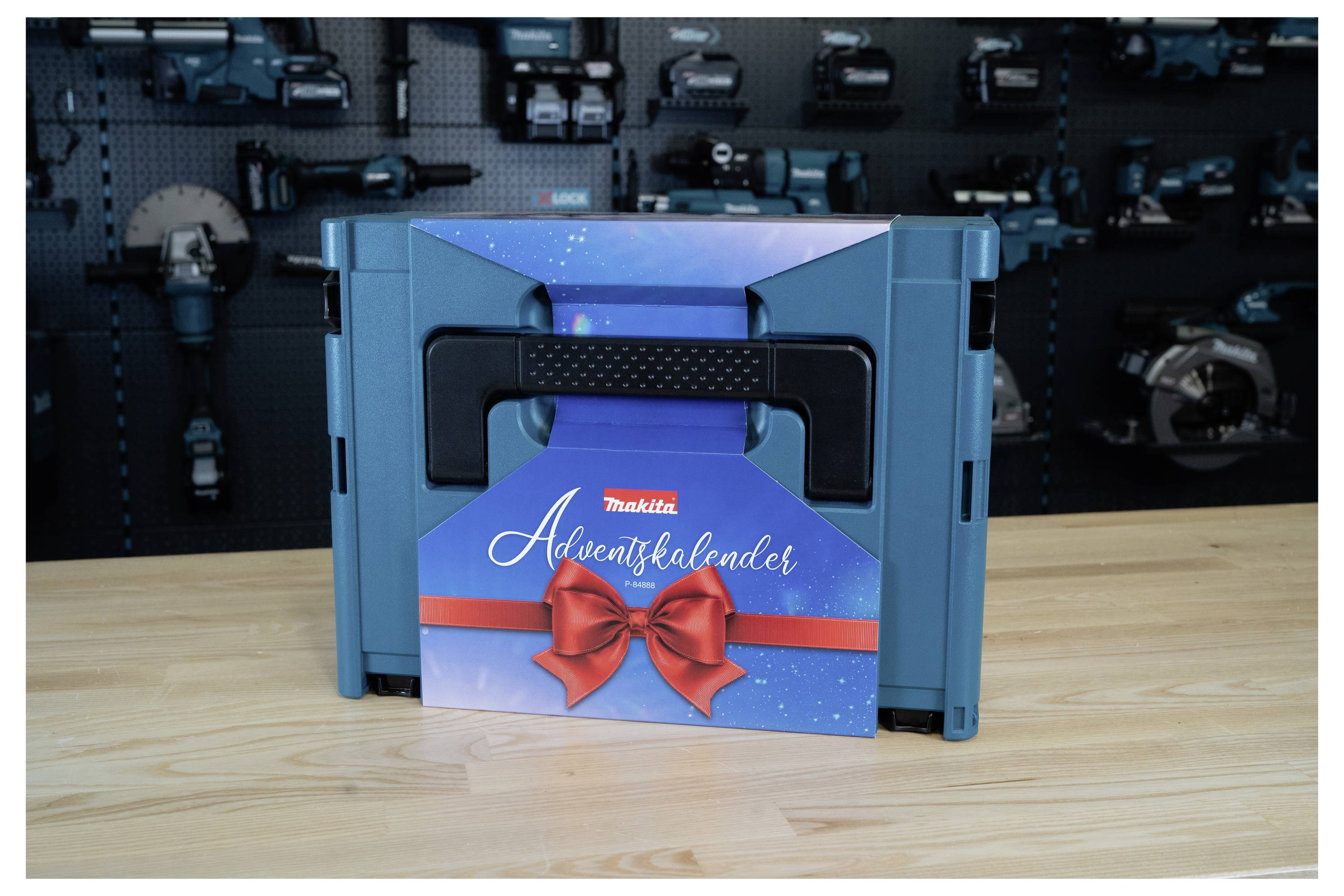 Blauer Makita Adventskalender in einer Geschenkbox mit rotem Band und Schleife auf einem Holztisch, vor einer Werkzeugausstellungswand.