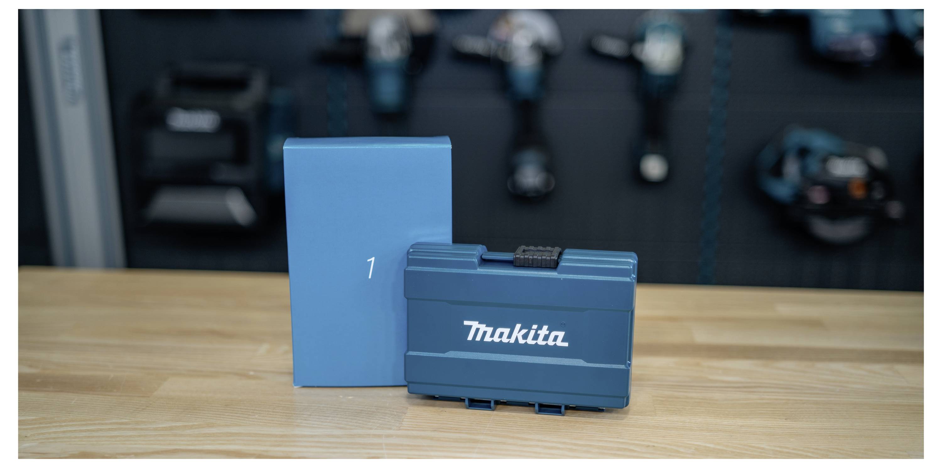 Blauer Makita-Werkzeugkoffer und passende Box auf einem Holztisch, mit unscharfen Elektrowerkzeugen im Hintergrund.